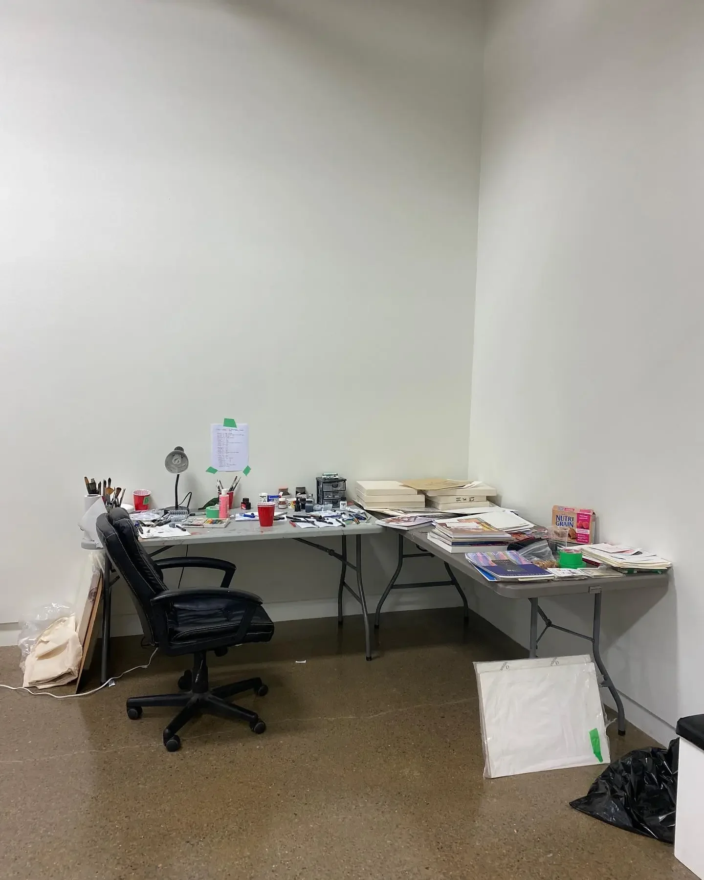 Disorganized office desk with scattered papers, books, office supplies, a black office chair, and some trash bags on a brown carpet.