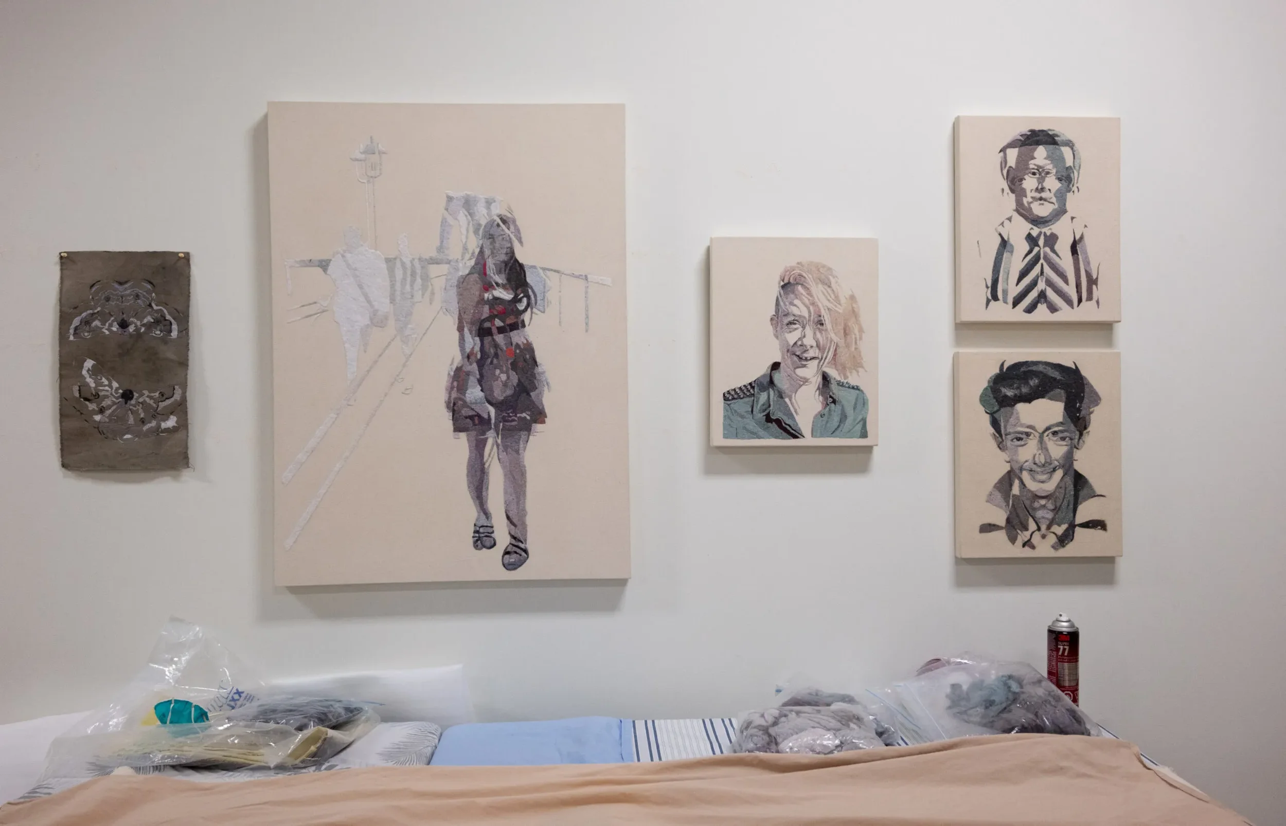 A wall decorated with six pieces of artwork, including portraits and abstract designs, above a bed with plastic bags and a can of spray paint on a side table.