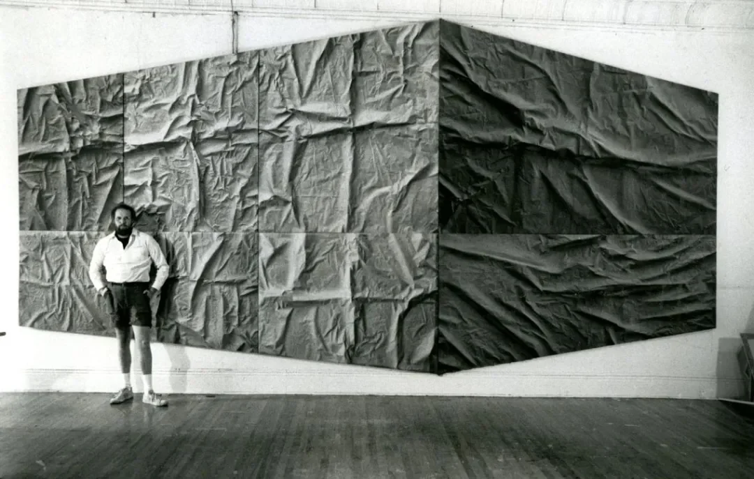 a man standing in front of a large scale multi panel art work in the space of a gallery.