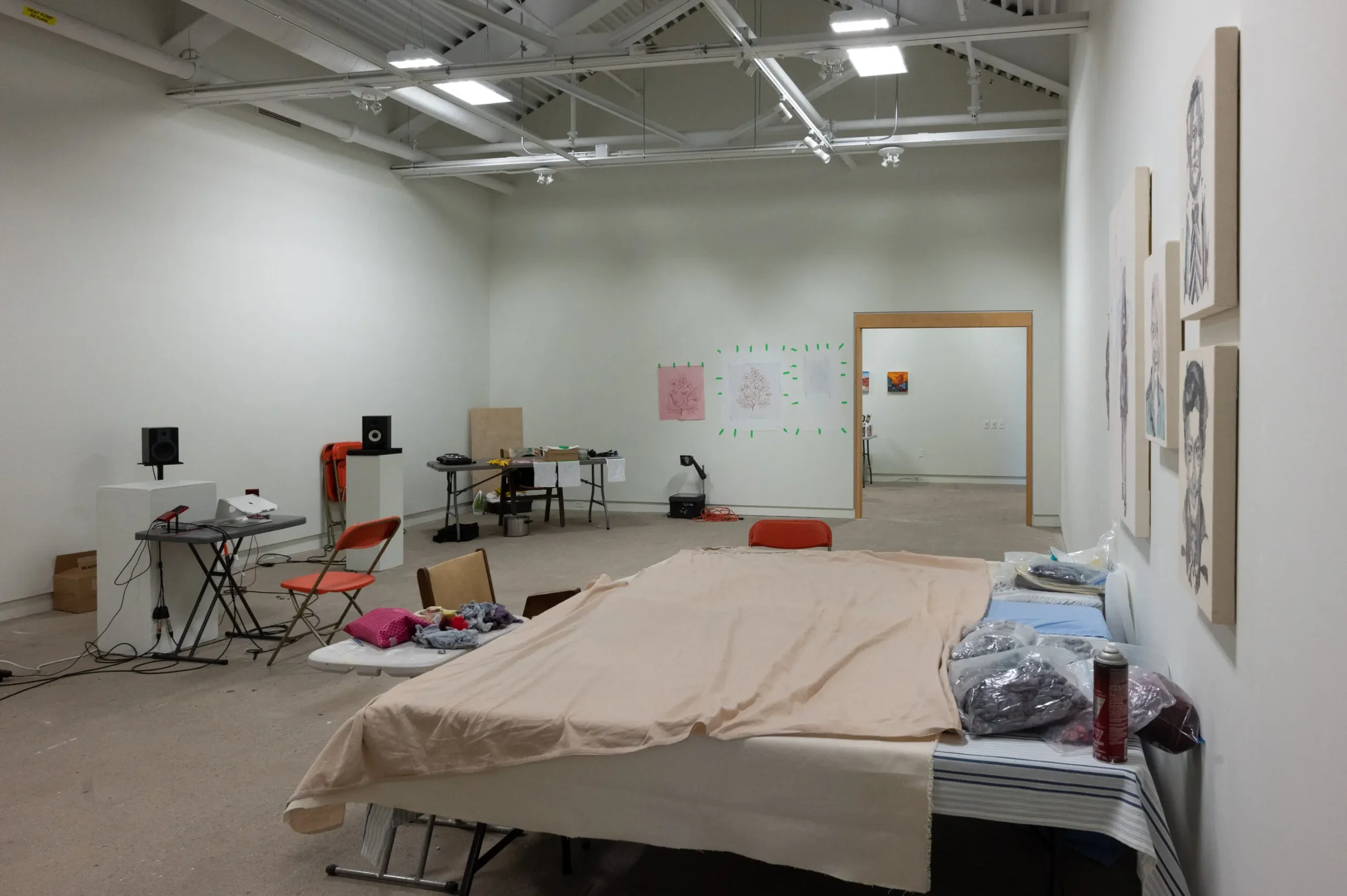 An art gallery or studio space with white walls, featuring artwork on the right wall and drawings on the back wall. There is a bed or examination table cluttered with clothes and items, along with chairs, tables, and sound equipment scattered through