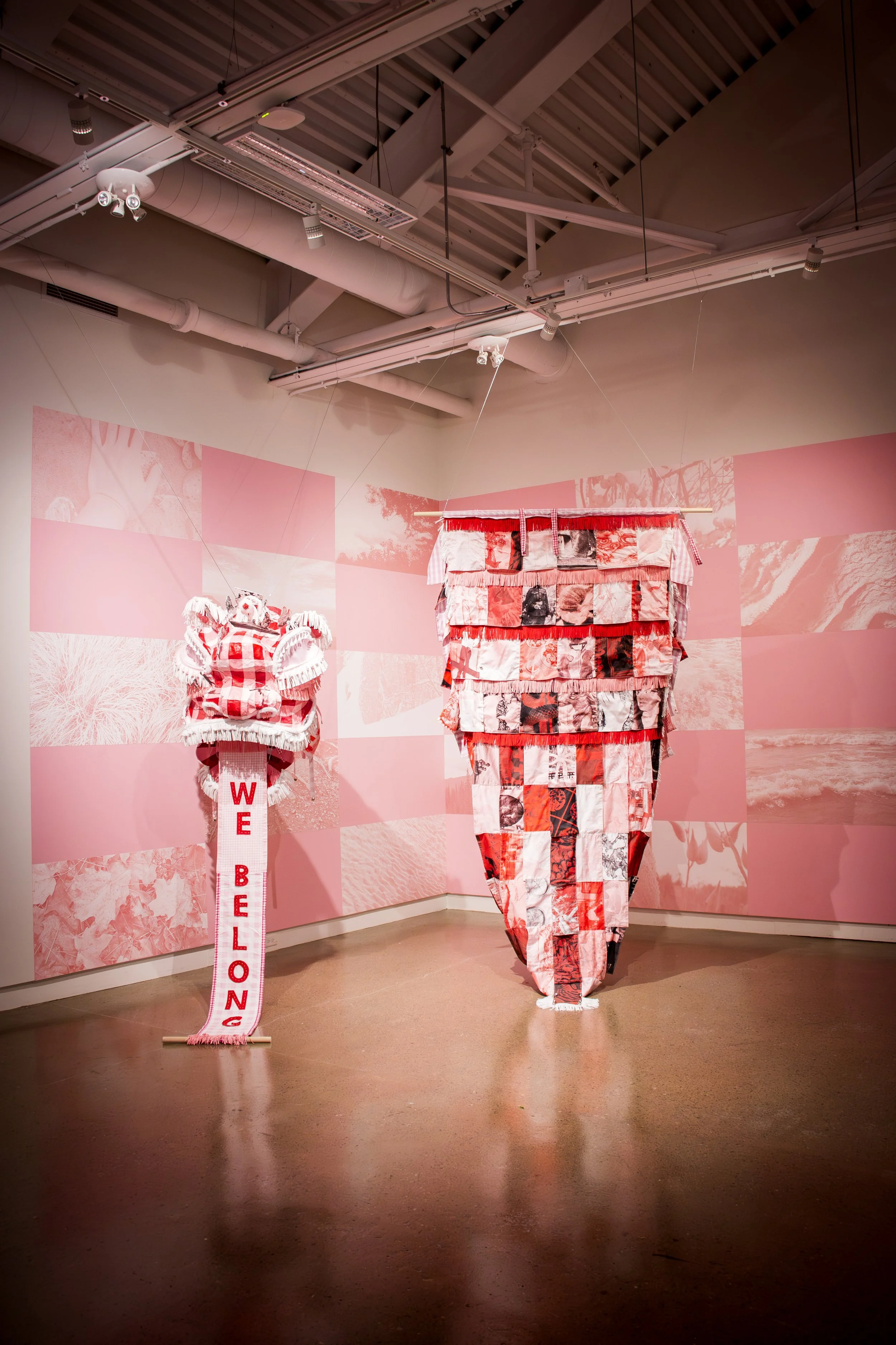 Art installation featuring two fabric sculptures, one with the words 'WE BELONG' in red letters on a white background, and the other made of patchwork fabric in red, black, and white, set against a pink-toned photographic background on the walls.