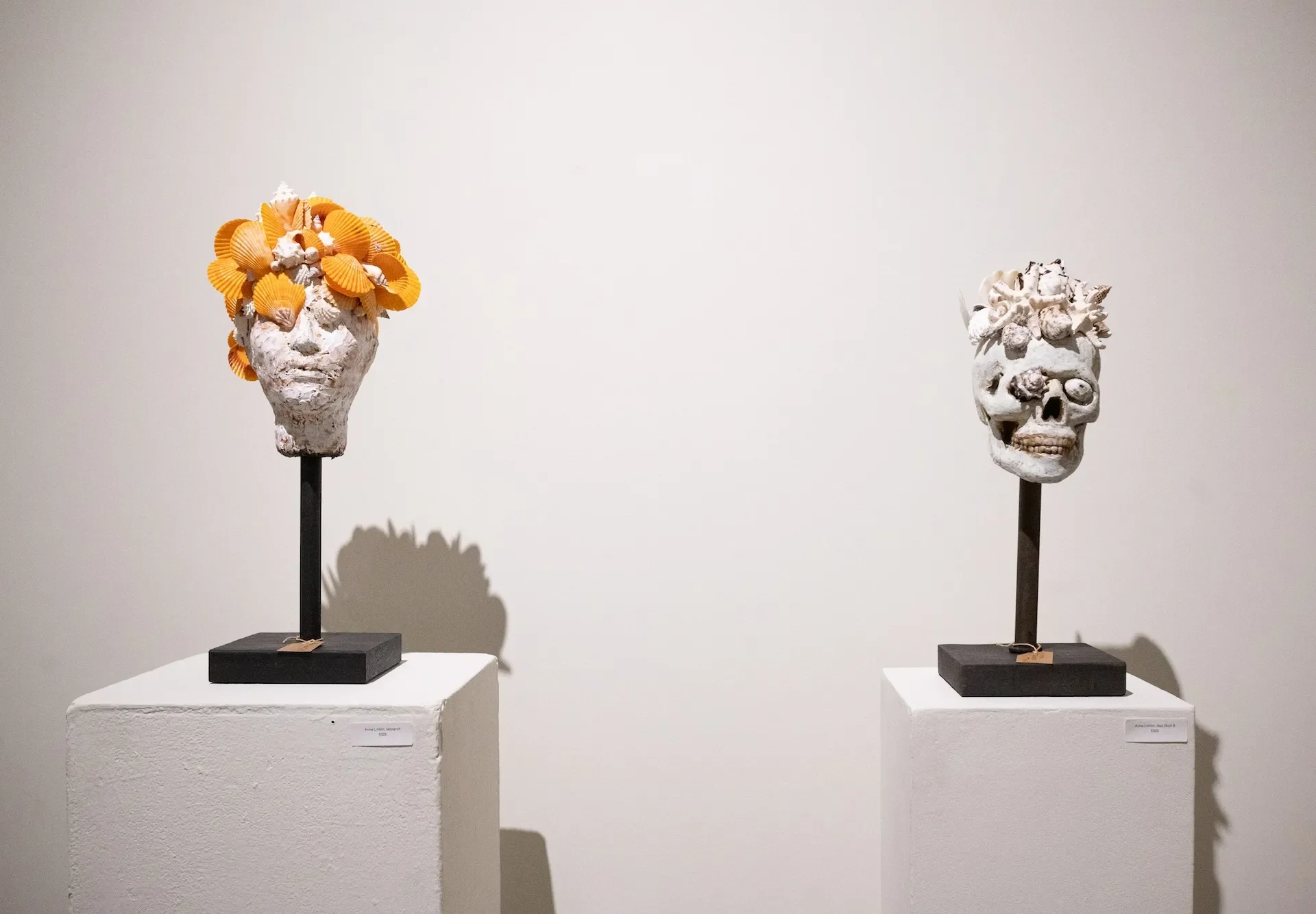 Two art sculptures of human heads on pedestals, decorated with seashells and other ocean elements, against a white gallery wall.