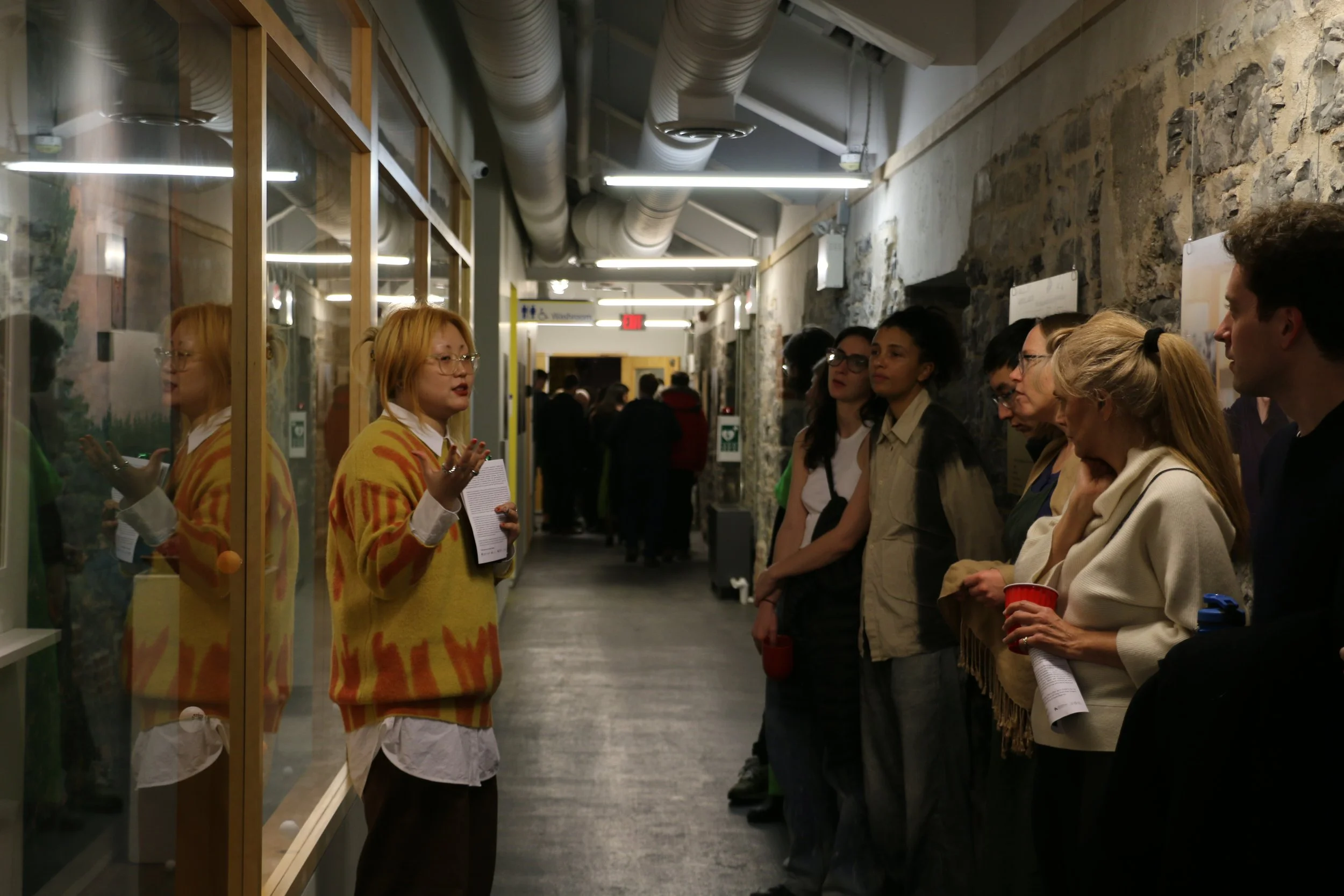 A woman with red hair and glasses is speaking to a group of people in a hallway with stone walls and industrial ceiling pipes.