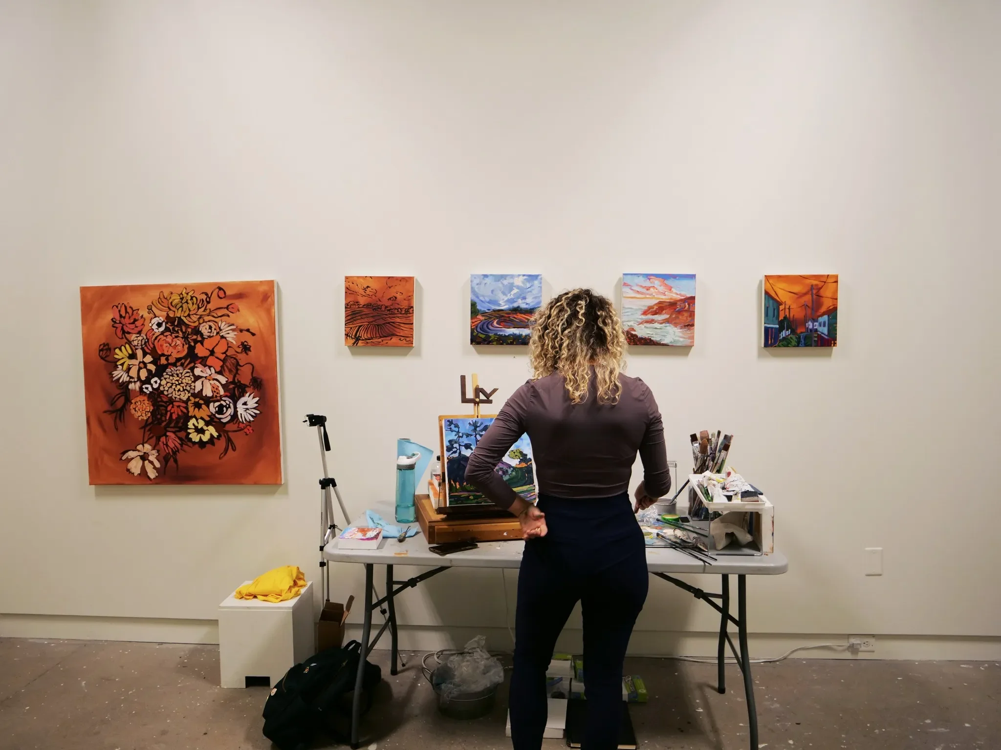 An artist standing at a work table in an art gallery, viewing five colorful landscape paintings on a white wall.