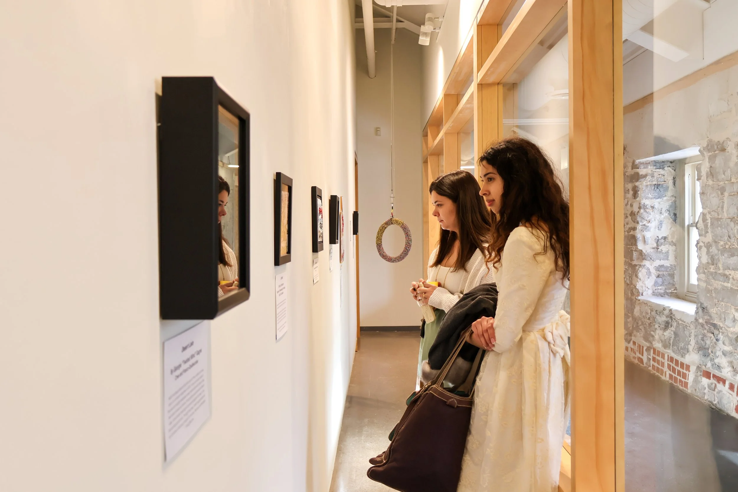 2 women on the inside of the Window Gallery looking at framed pieces on the wall.
