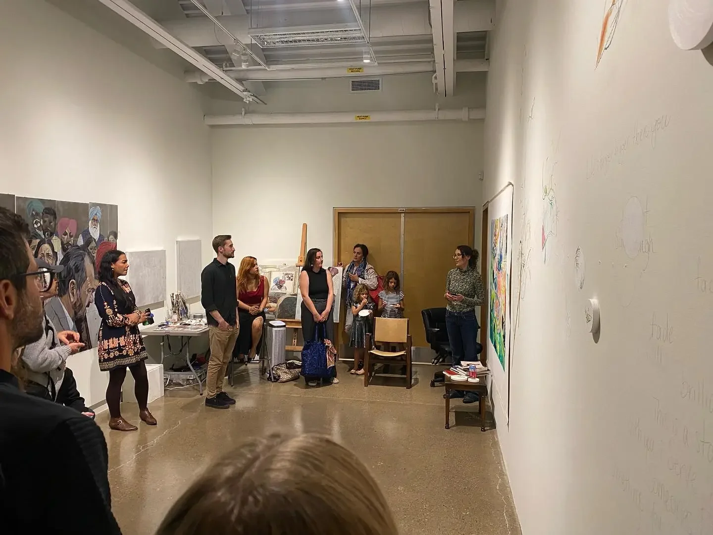 A woman giving a presentation or lecture to a group of people in an art gallery or classroom, with artwork on the walls and children present.