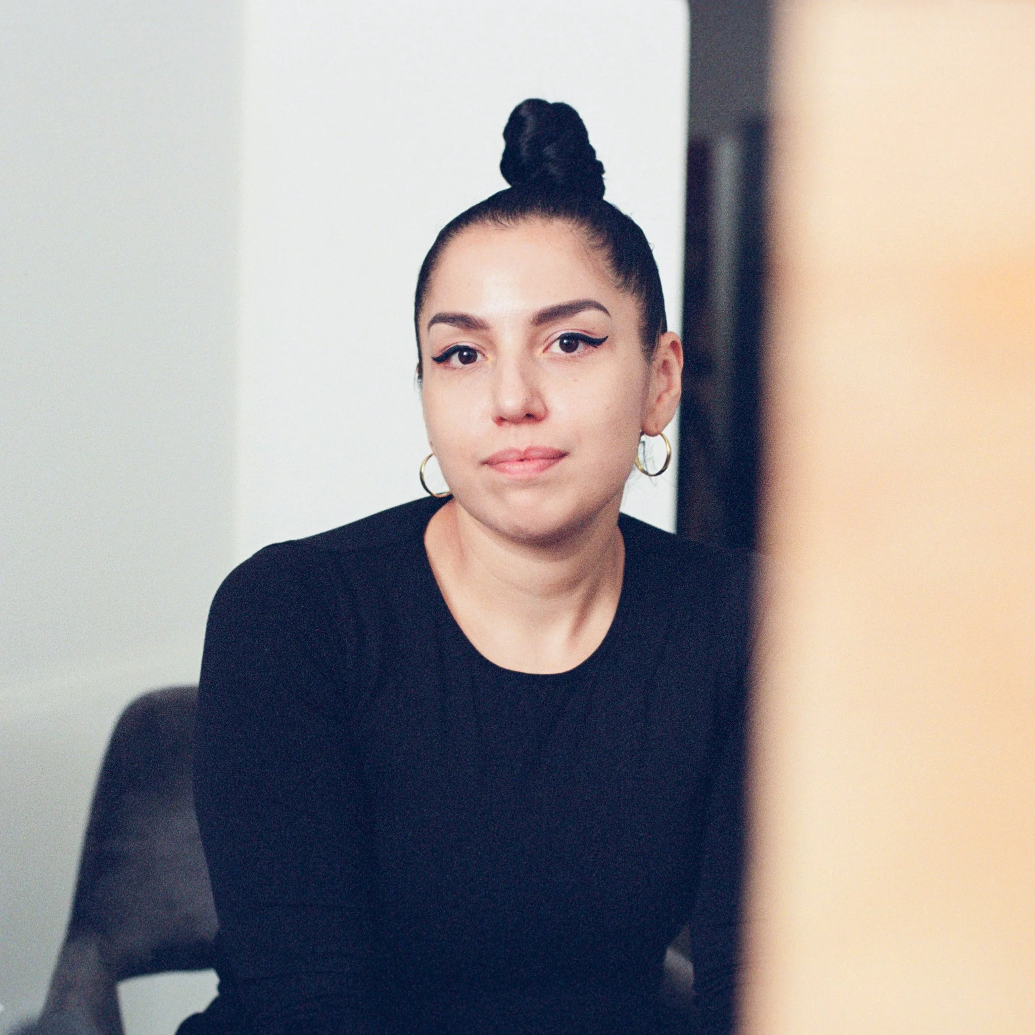 Young woman with dark hair in a high bun, wearing hoop earrings and a black shirt, looking at a computer screen with a neutral expression.