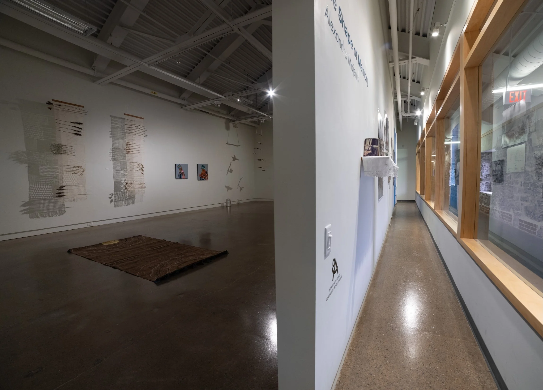 View of two art gallery sections separated by a wall. The left section features textile wall hangings and artwork, and a woven rug on the floor. The right section shows a hallway with framed art pieces and a large window with wooden framing, allowing natural light.