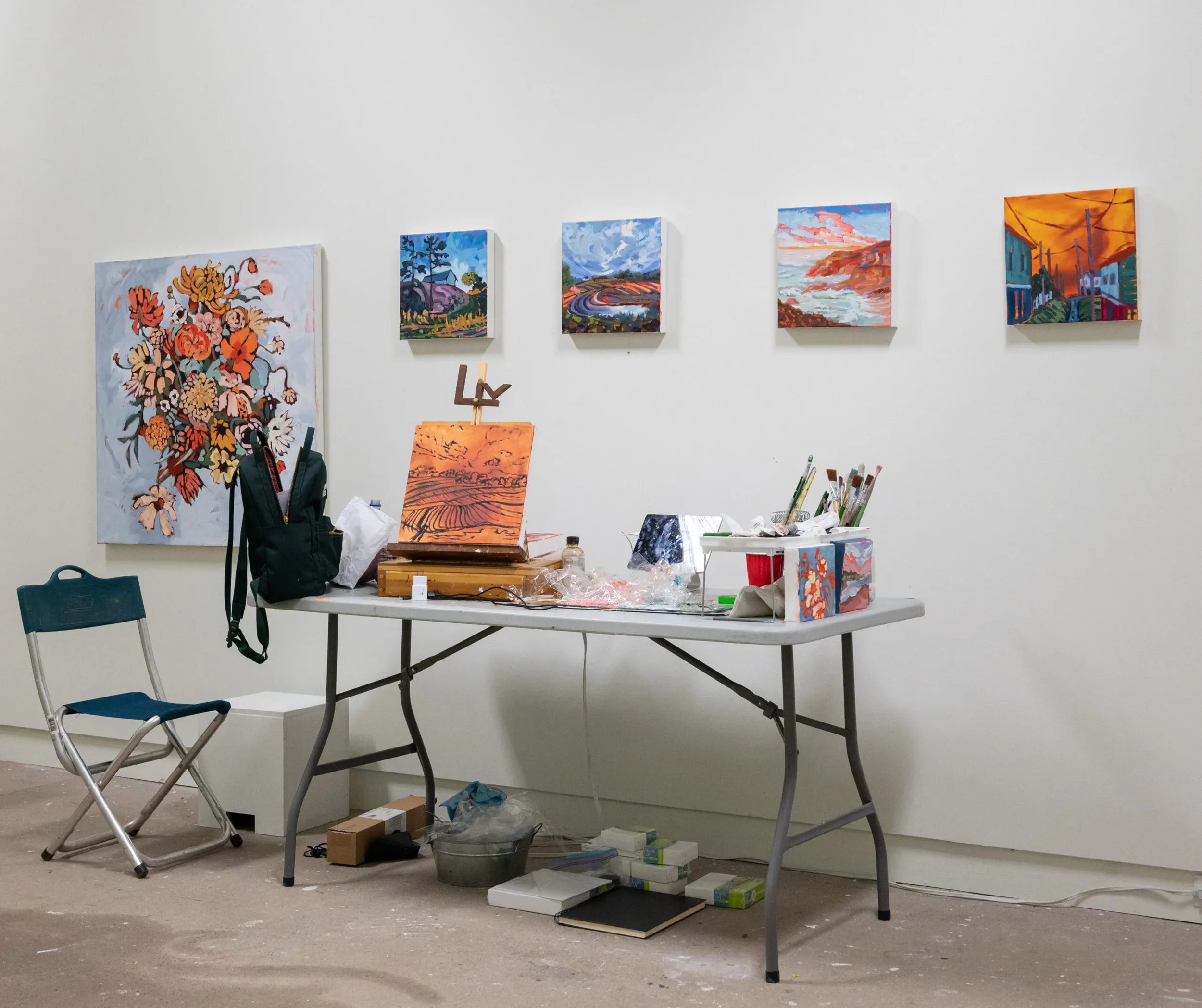 An art studio with a white wall displaying five colorful landscape paintings. A table with painting supplies, including brushes and paints, is in front of the wall. There is a black backpack on the table, and a blue folding chair to the left.