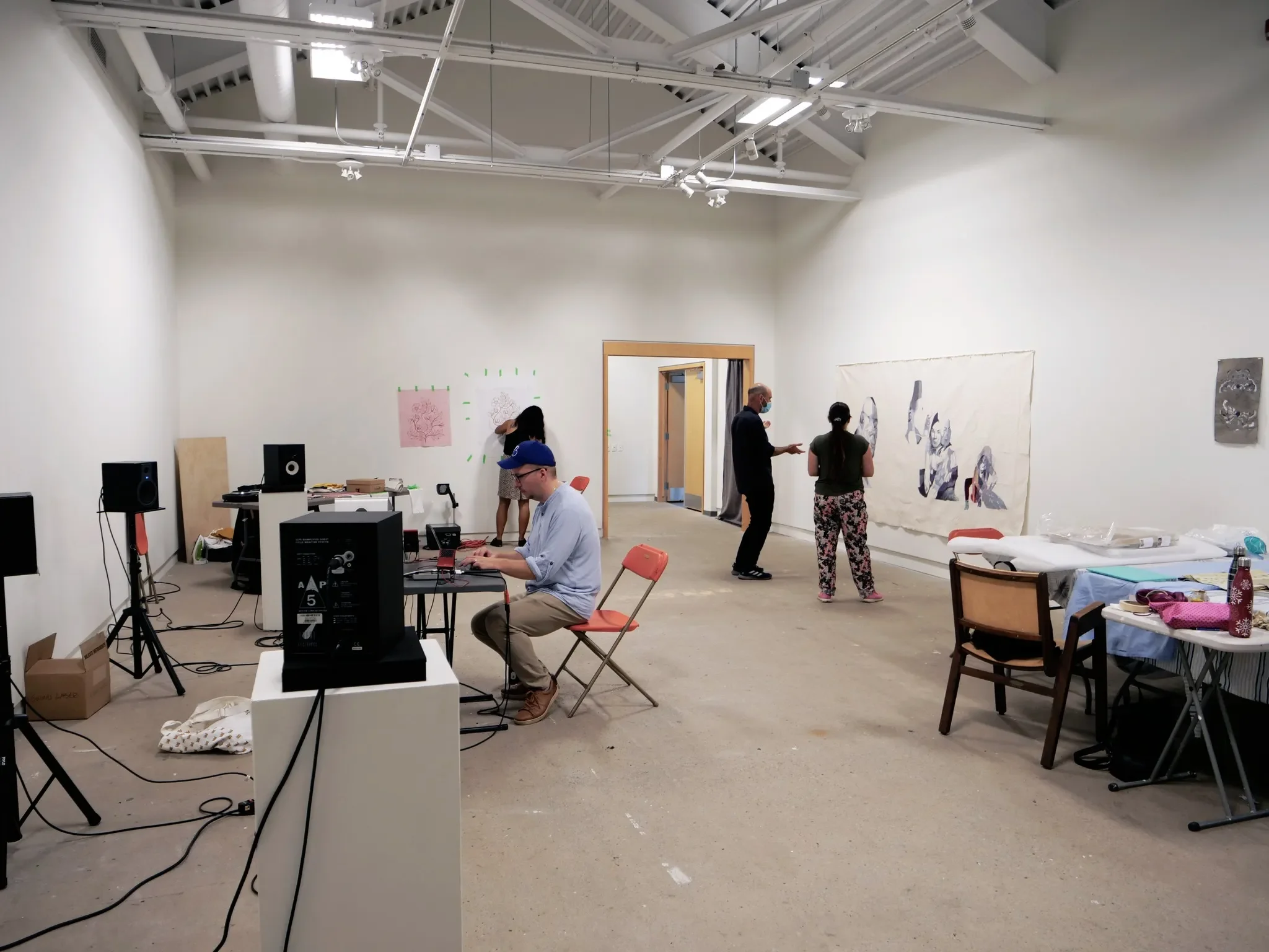 People in an art gallery or studio, some working on a computer, others discussing large artwork on the wall, and one person arranging art supplies or fabric on tables.