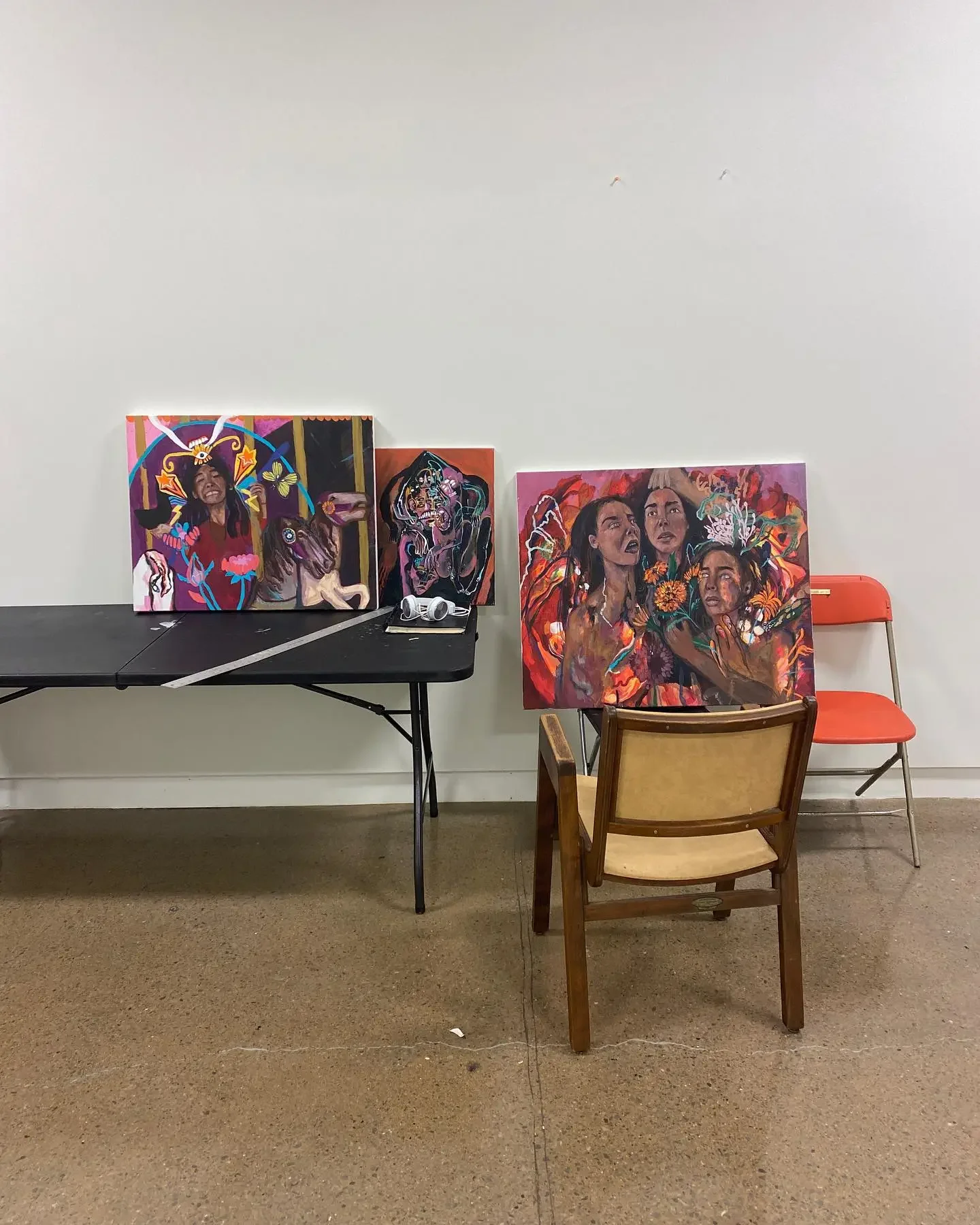 Table with three art paintings leaning against a plain white wall, beside two chairs, one red and one with a wooden frame and beige seat, on a brown carpeted floor.