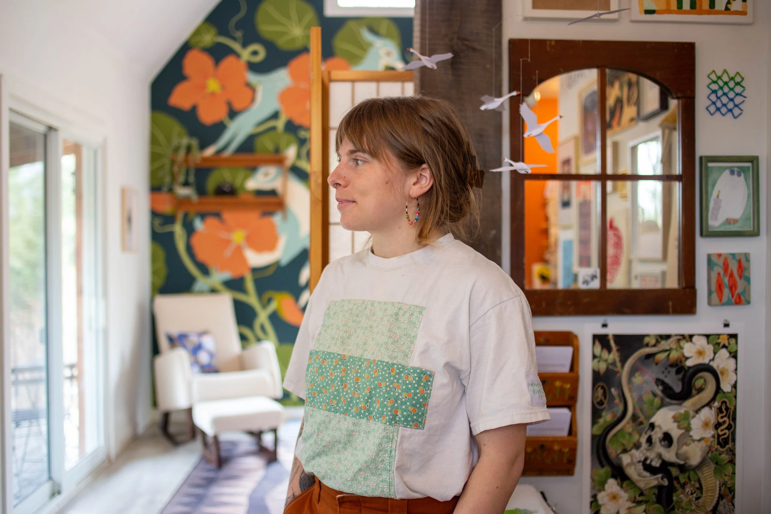 A woman with short brown hair and colorful hoop earrings standing indoors, with colorful artwork and hanging paper birds in the background.