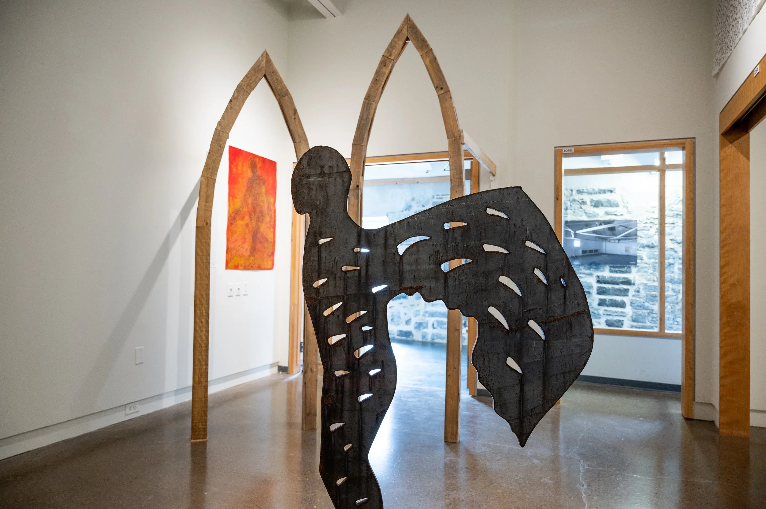 An art gallery display featuring a wooden silhouette sculpture of a human head and torso in front of a church-like wooden arch, with paintings on the walls and a window showing a stone wall outside.