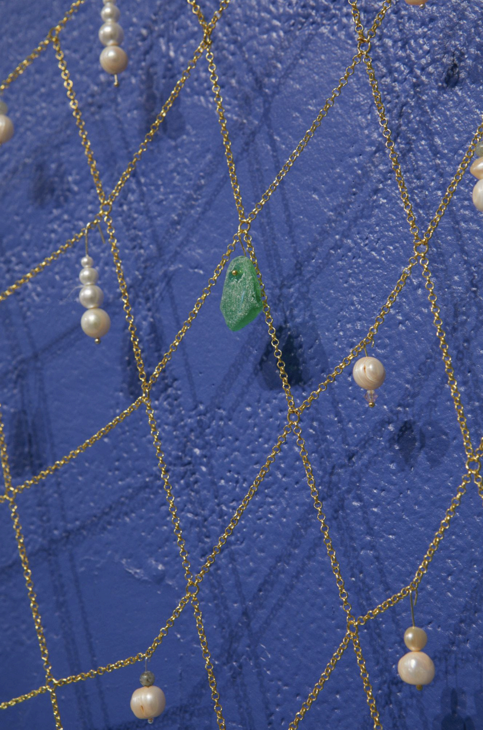 Gold chain necklaces with pearls and a green stone pendant hanging on a textured blue wall.