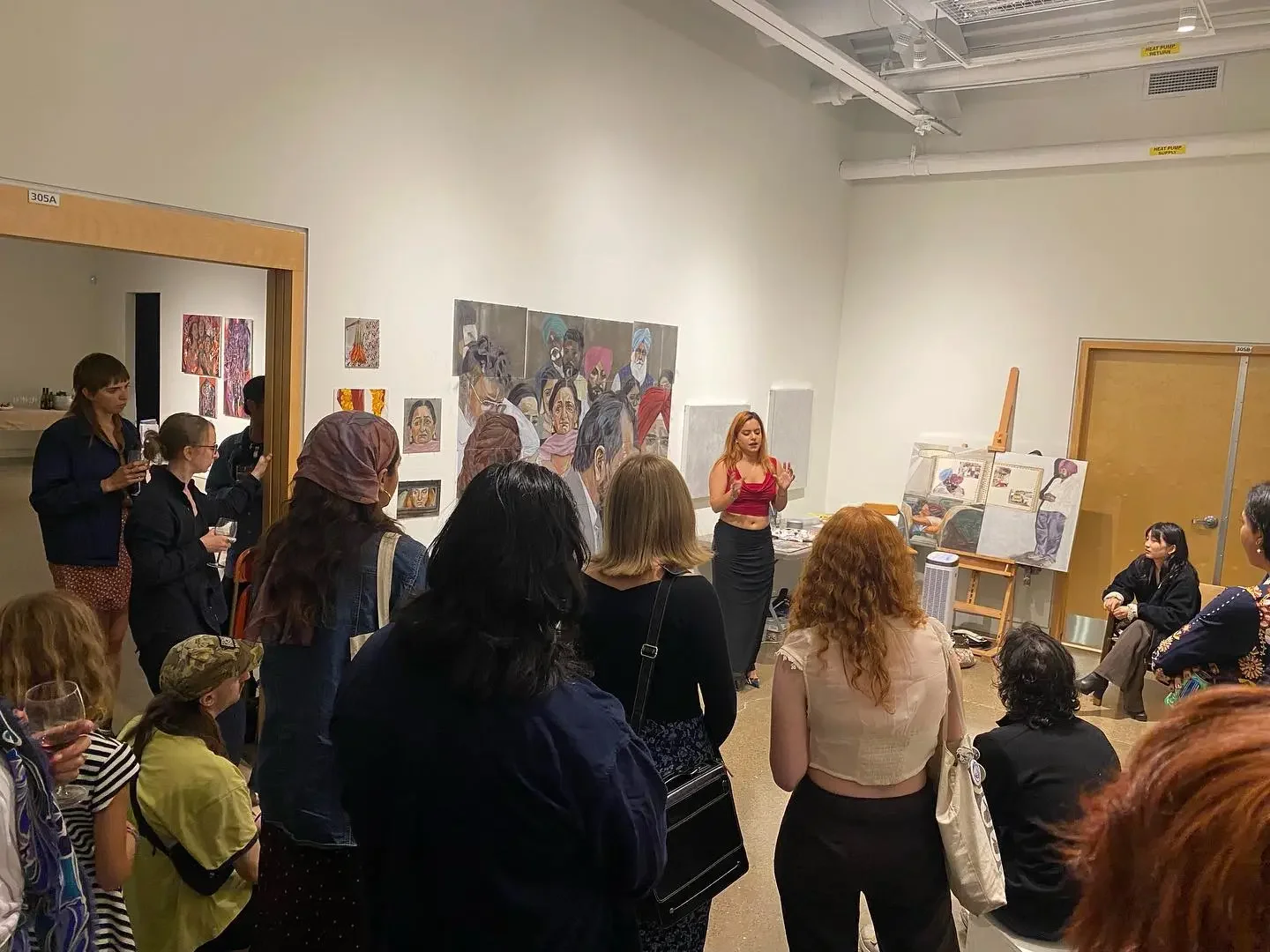 An art gallery with a group of people gathered around a female artist giving a talk. The walls display various colorful portraits and artworks, and an easel with paintings is visible on the right.