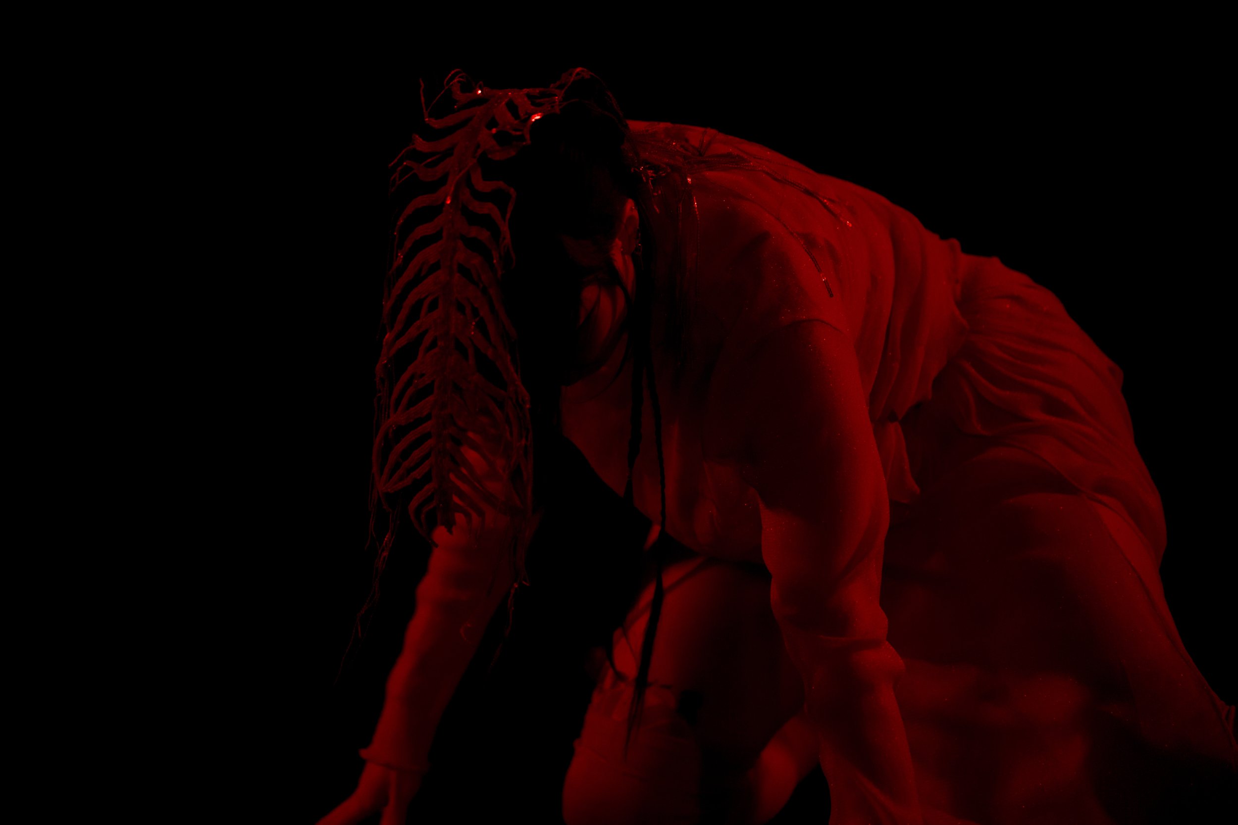 A person with dreadlocks and a muscular physique, covered in red lighting, standing against a black background.