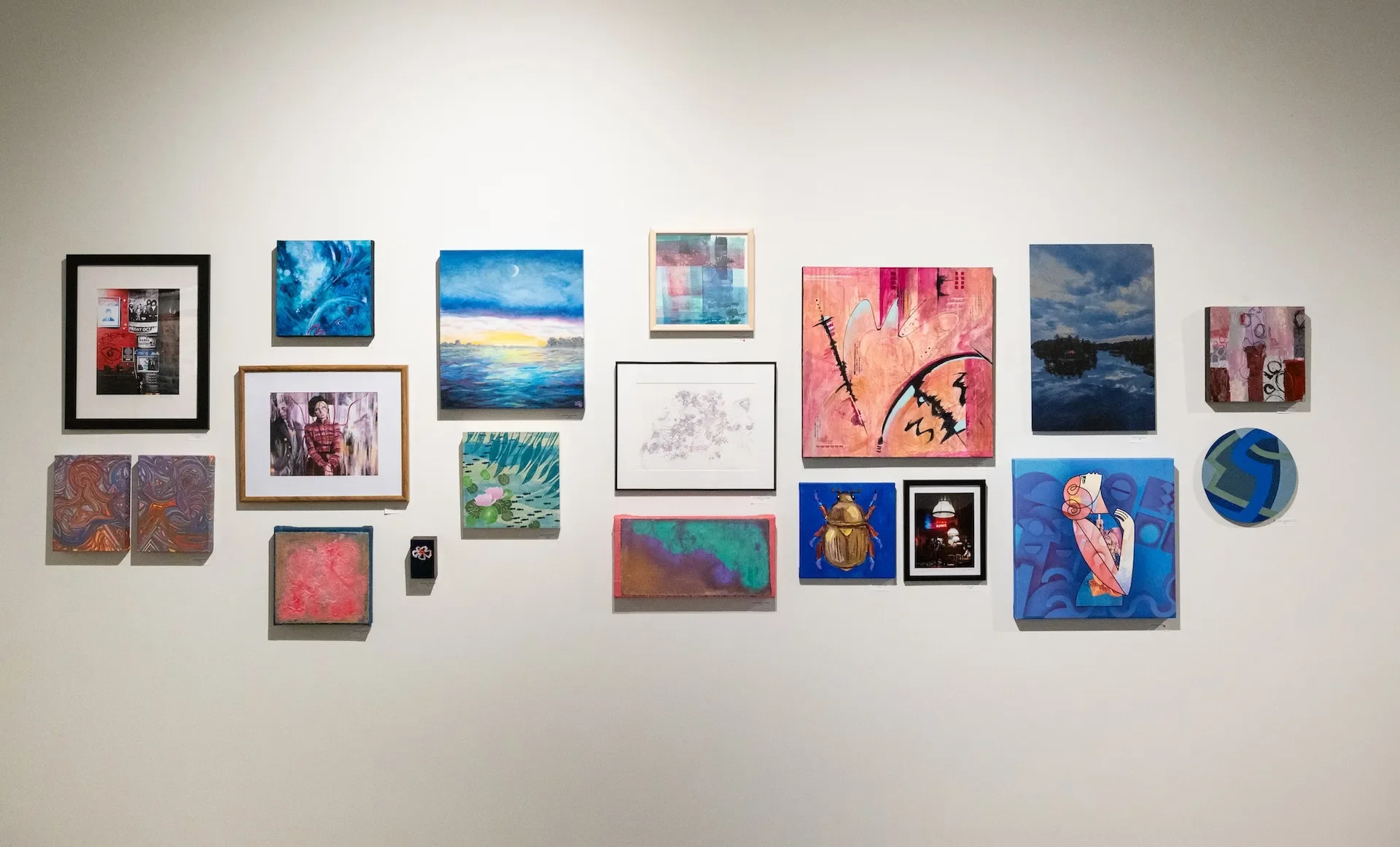 A white gallery wall displaying a diverse collection of framed and unframed artwork, including paintings, photographs, and mixed media pieces in various styles and colors.