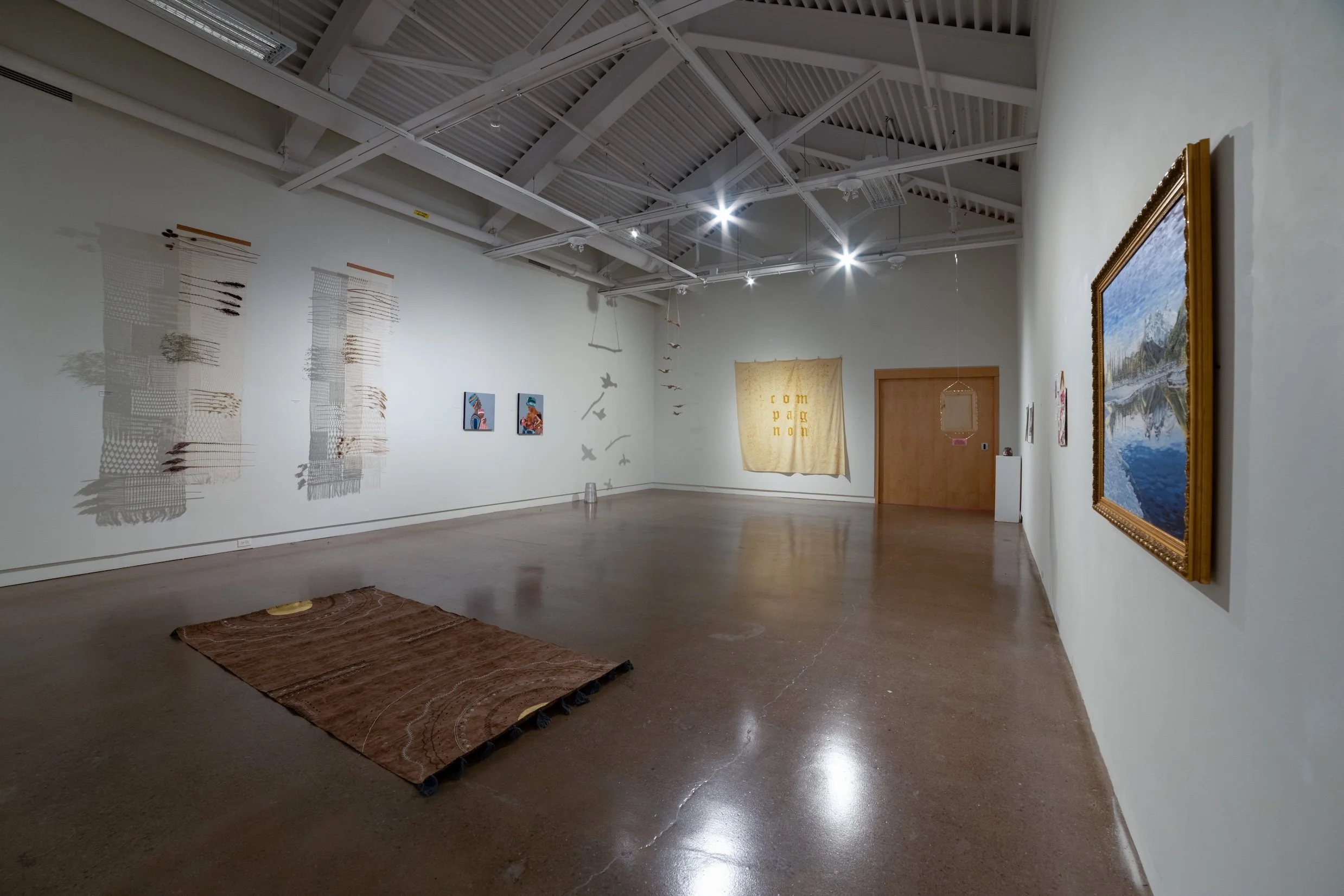 Interior view of an art gallery with various artworks including paintings, textiles, and mixed media pieces on white walls, with a polished concrete floor.