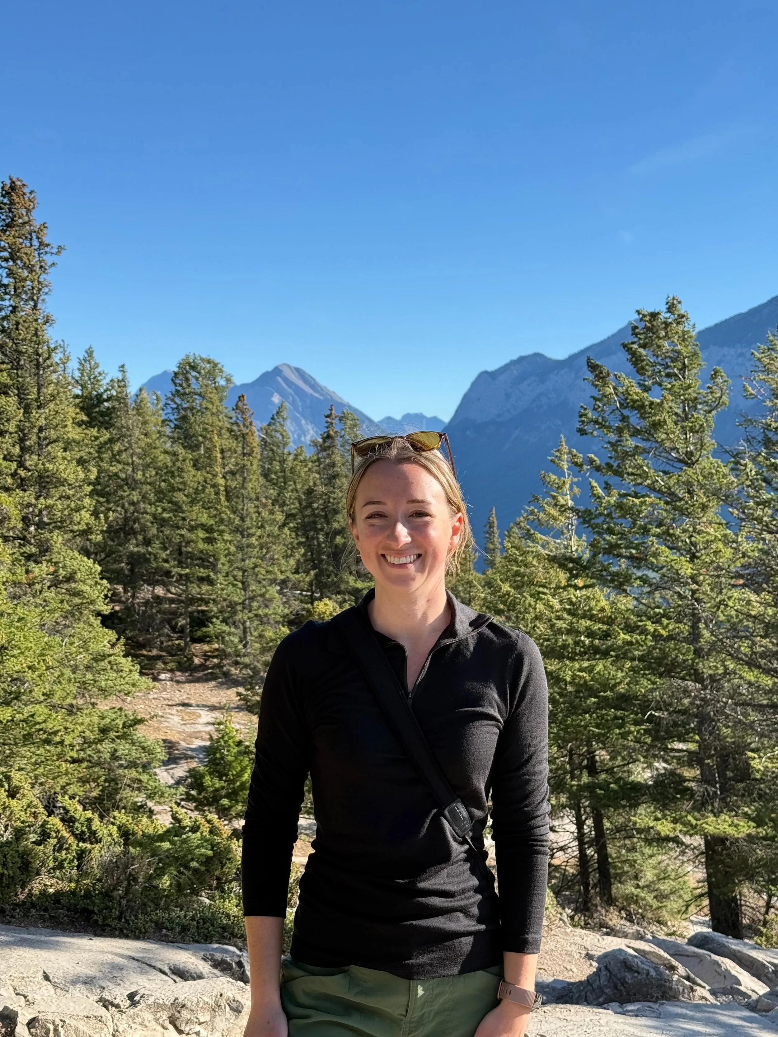 A woman smiling outdoors in a forested mountain area with blue sky and mountains in the background.