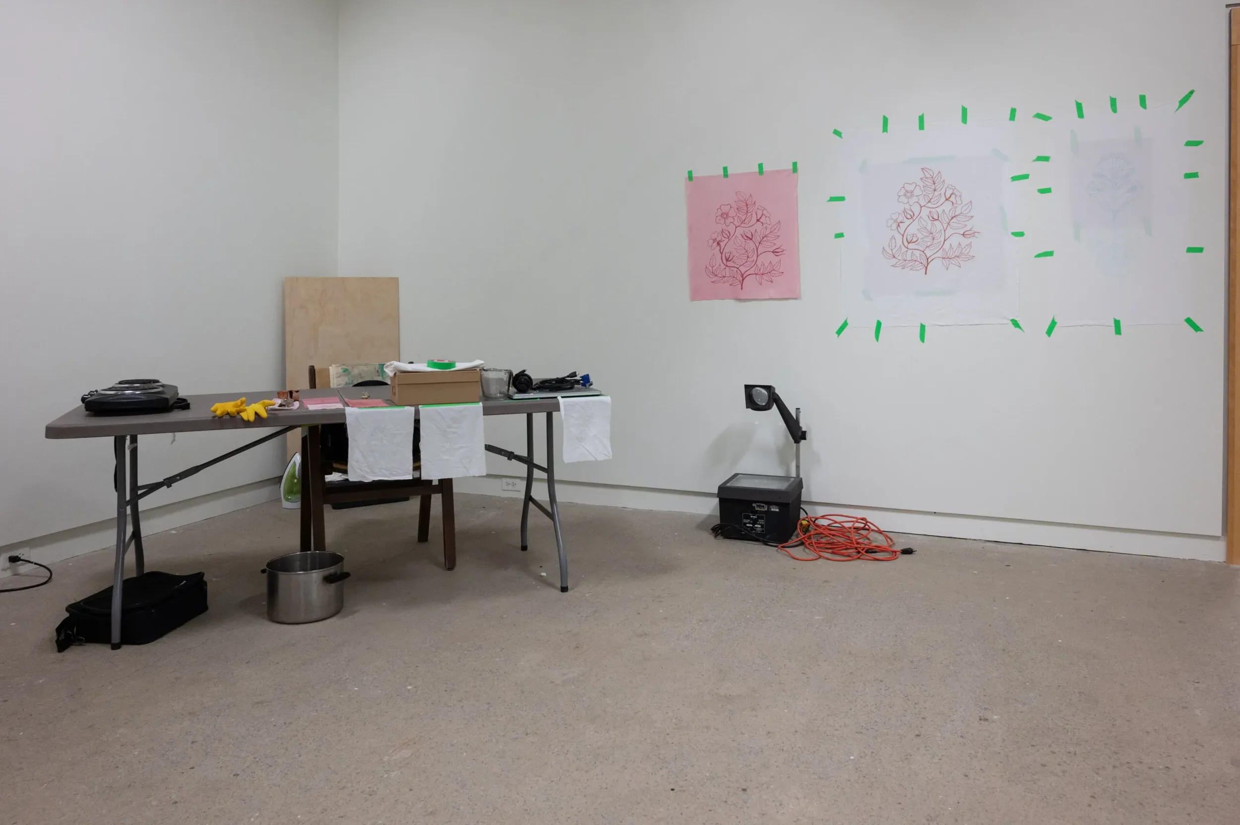 An empty room with a table, arts and crafts supplies, and three sketches of plants taped to the wall with green tape.