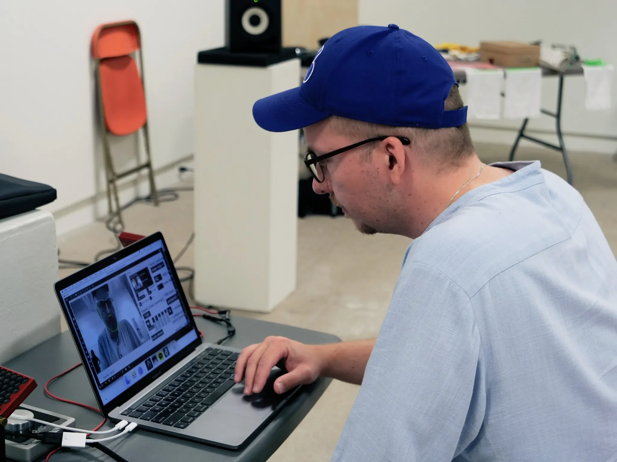 A man sitting at a desk working on a laptop with a video call displayed on the screen, wearing glasses, a blue baseball cap, and a light blue shirt.