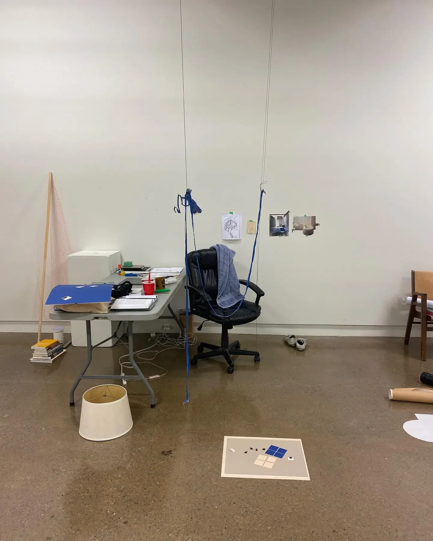 An office with a cluttered desk, a black office chair with a gray jacket, a swing hanging from the ceiling, and miscellaneous items on the floor and walls.