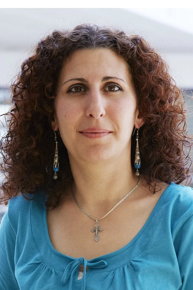 A woman with curly reddish-brown hair, wearing a turquoise top, silver cross necklace, and dangling earrings with blue beads, posing outdoors.