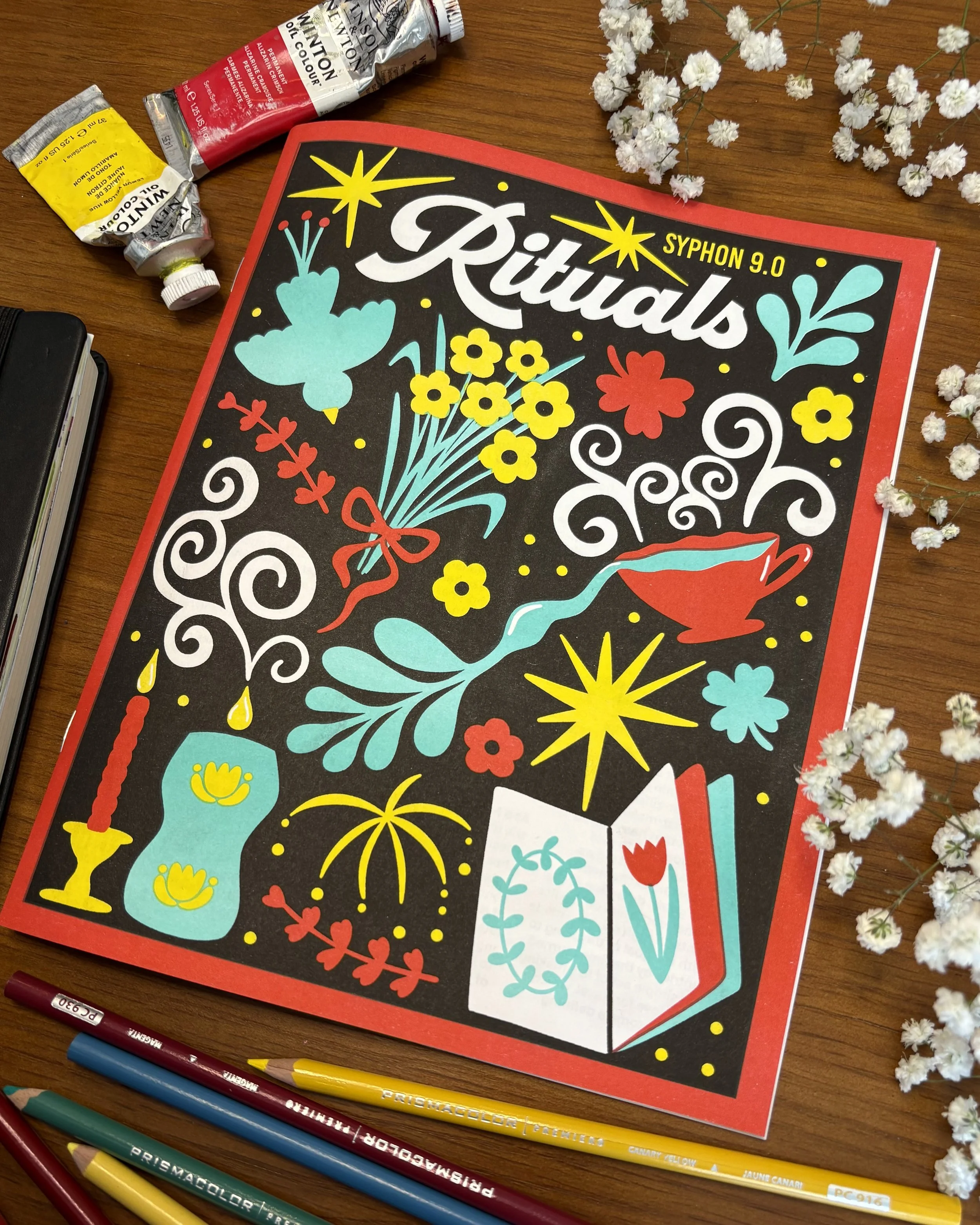 A colorful book titled 'Rituals' with illustrations of flowers, a butterfly, a candle, a book, and a cup, placed on a wooden table surrounded by colored pencils, small white flowers, and tubes of paint.