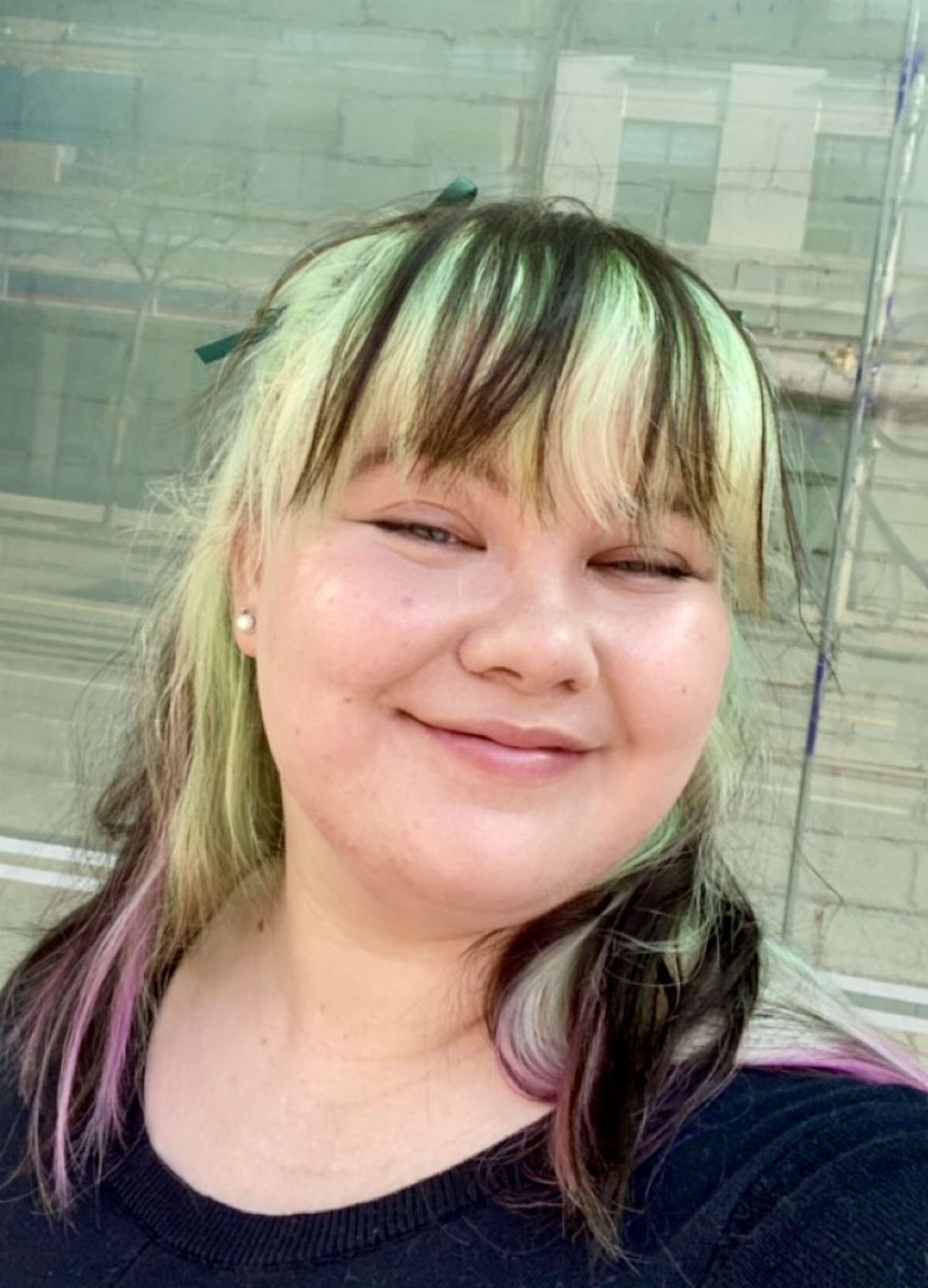 A woman with shoulder-length hair with green, blonde, and black streaks, smiling outdoors in front of a glass building.