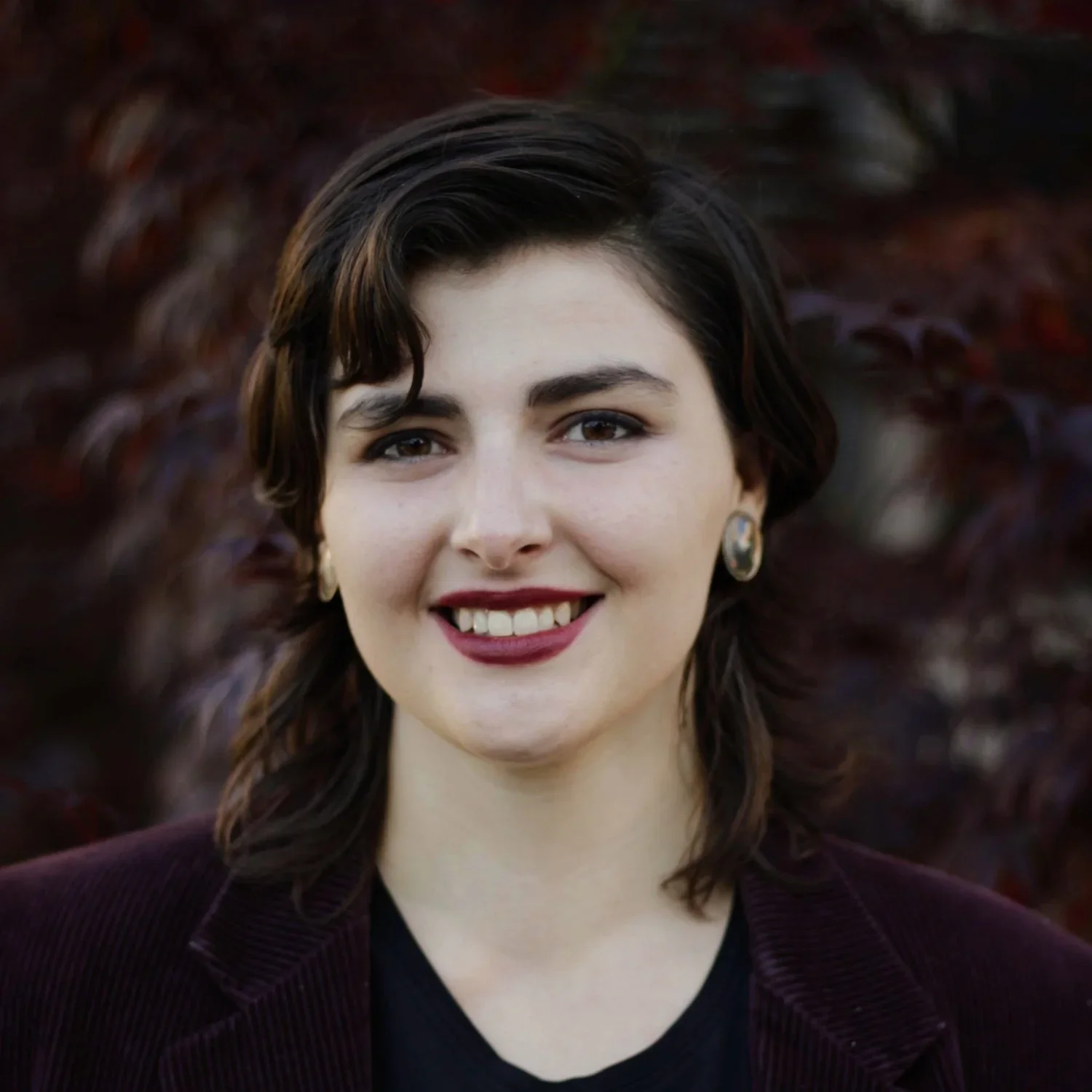 Close-up of a young woman with short dark wavy hair, wearing a dark top and earrings, smiling outdoors with blurred reddish-brown foliage in the background.