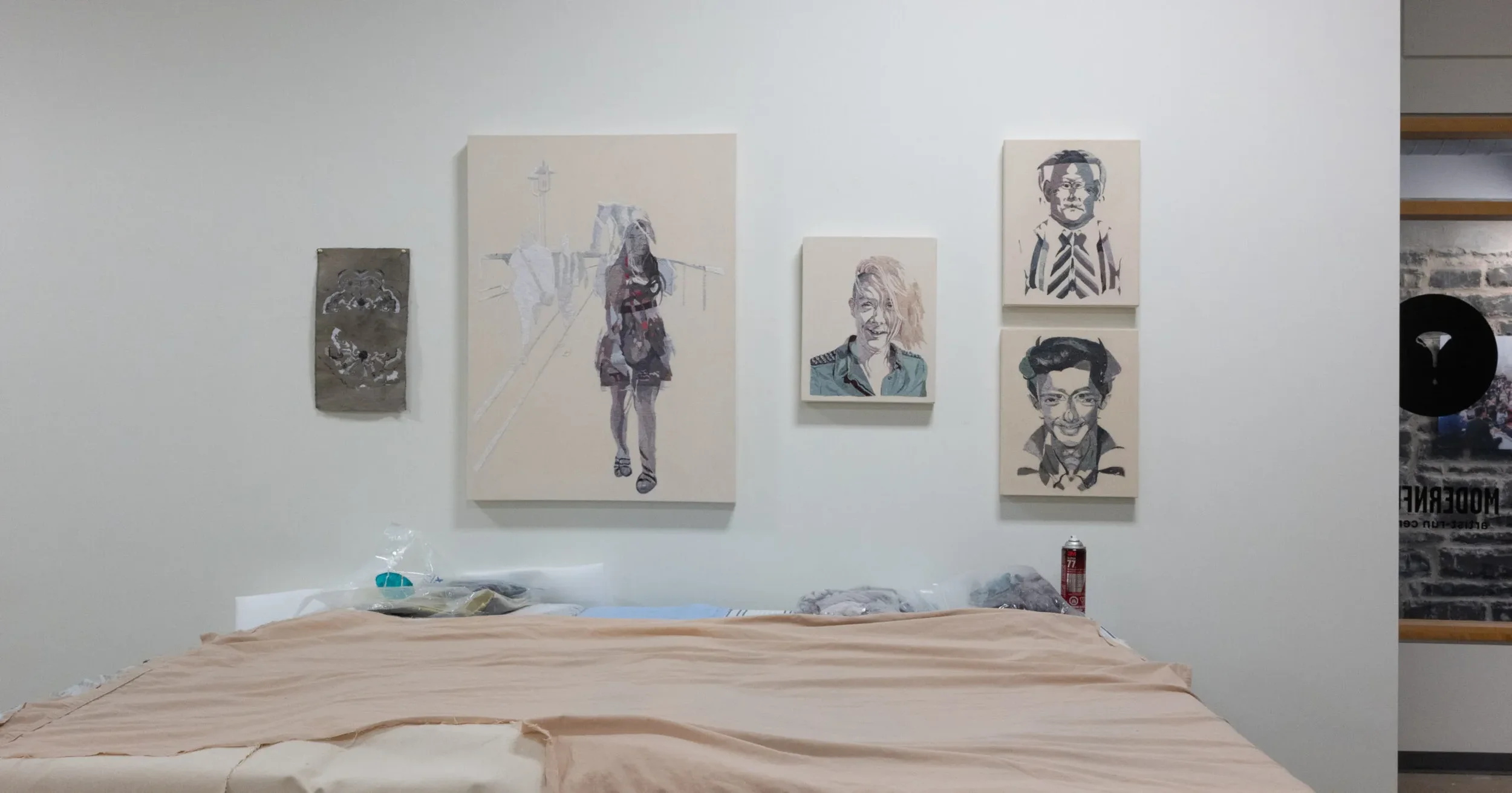 Gallery wall with five portraits of smiling individuals, and a woman walking on a bridge painted on canvas in the center. A bed with a beige sheet is in the foreground, with various items and a fire extinguisher visible on the side.