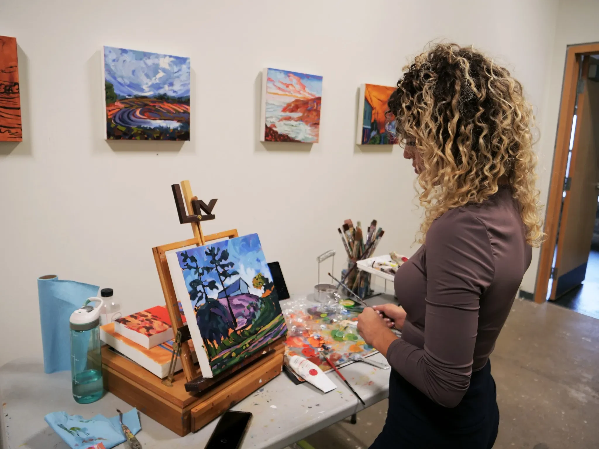 A woman with curly blonde hair painting a landscape on a small canvas in an art studio, with colorful landscape artworks hanging on the wall behind her.