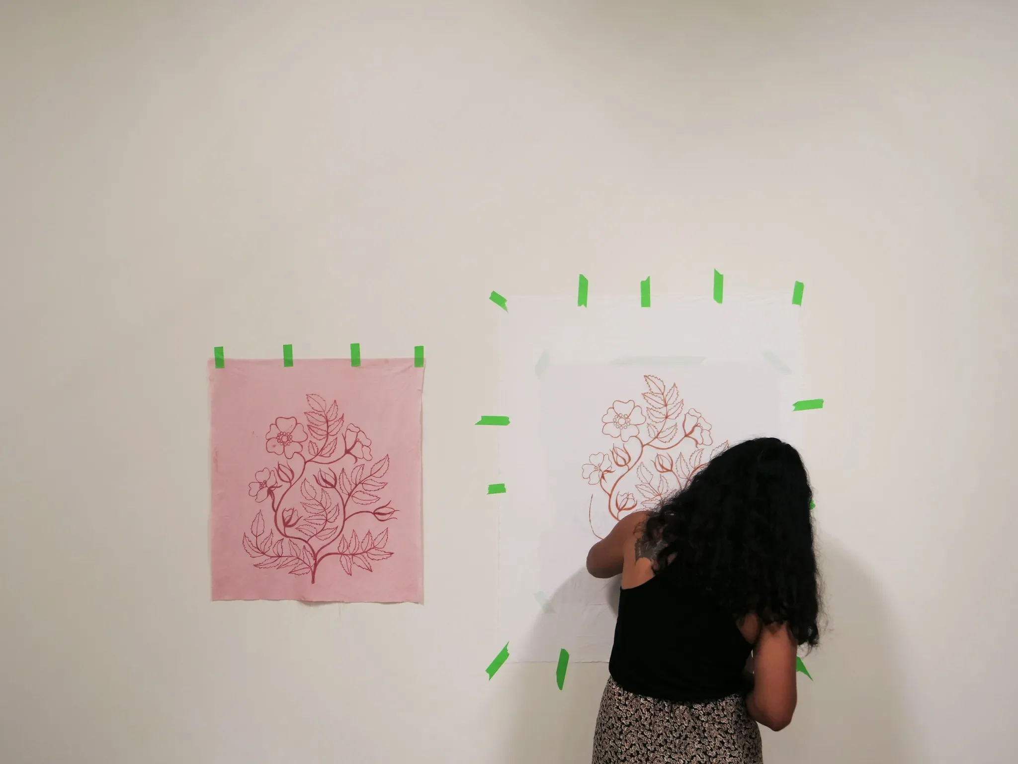 A woman with curly black hair, wearing a black sleeveless top and patterned skirt, is drawing a floral design on a large piece of paper taped to a white wall. There is a second floral drawing on pink paper taped beside it.