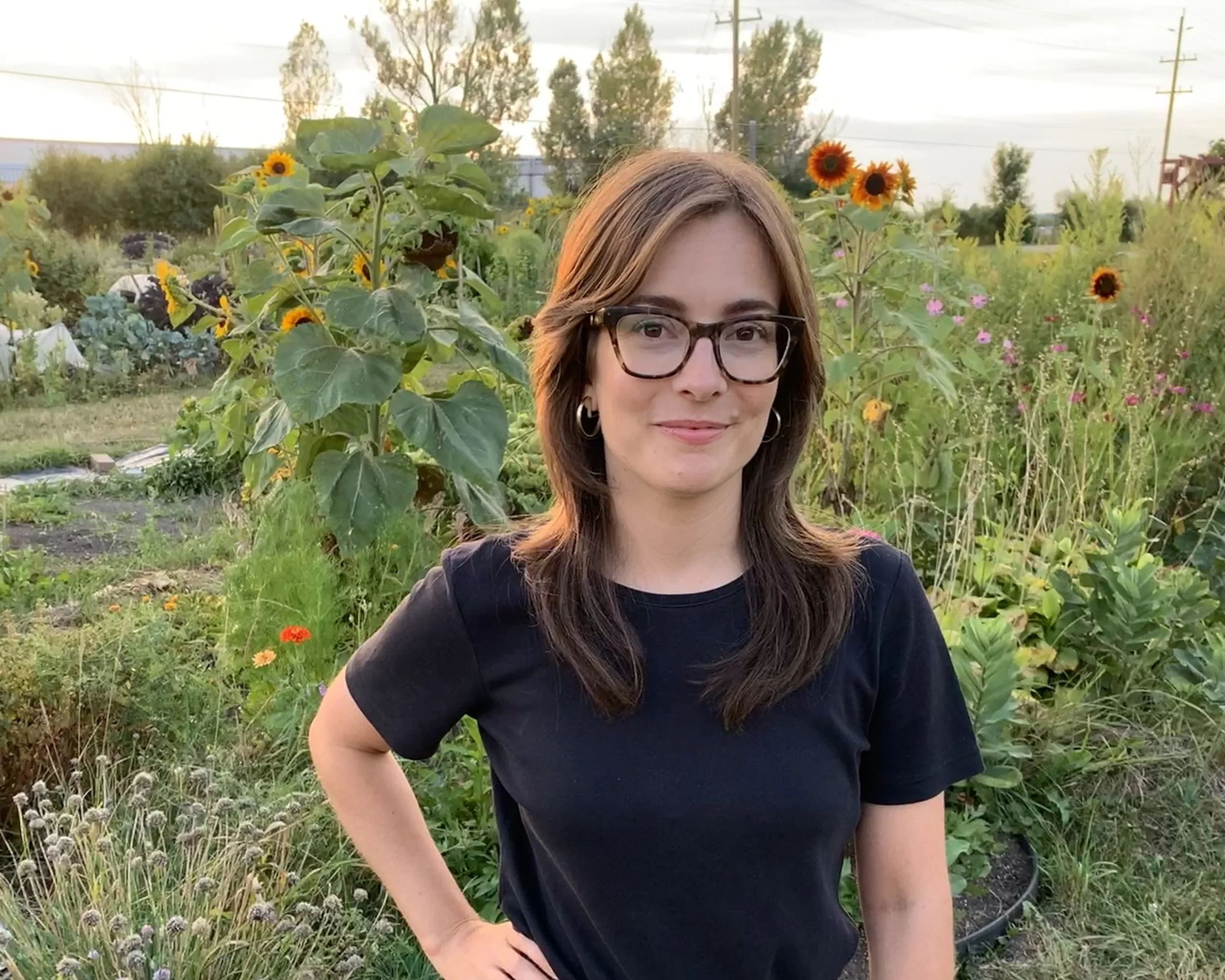 A woman with glasses and long brown hair standing in a garden with sunflowers and other flowers during sunset.