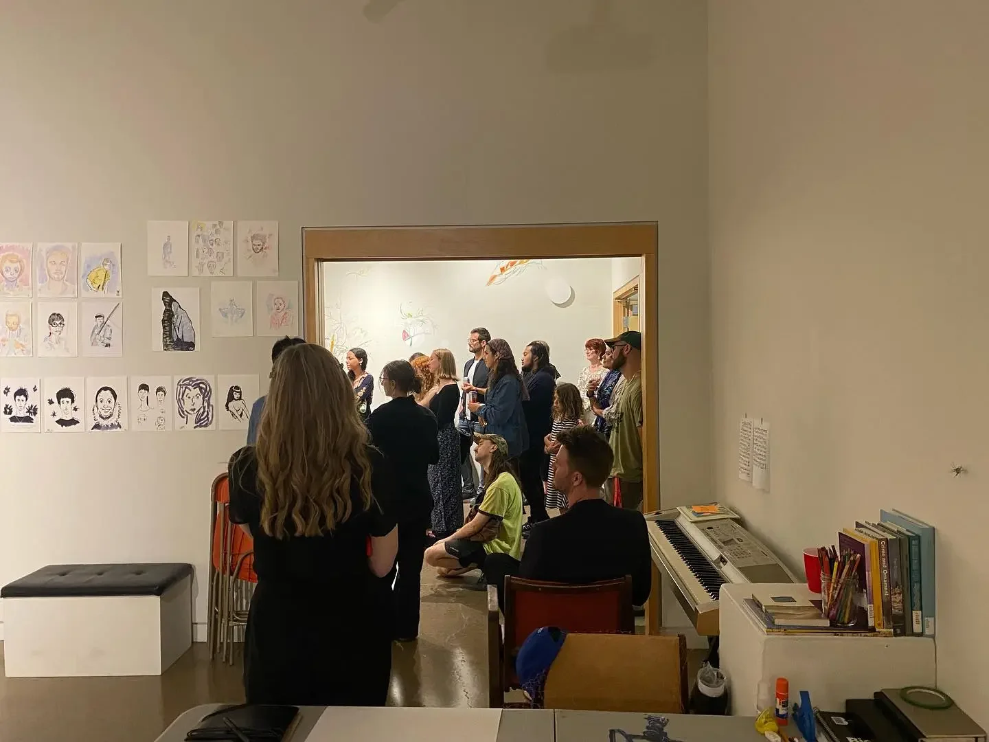 People attending an art gallery exhibition, with artwork displayed on the wall and a wood-framed doorway leading to another room.