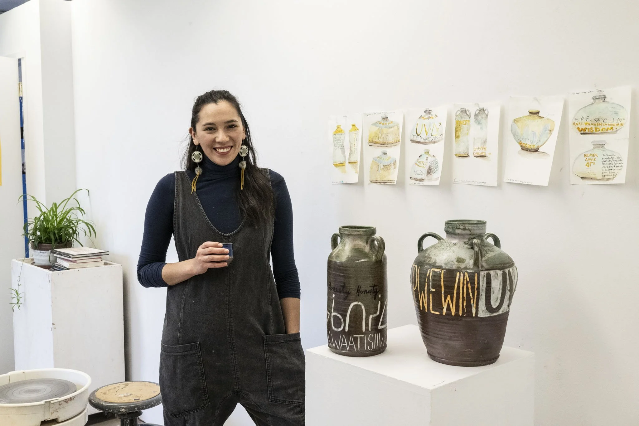 A young woman with long dark hair, wearing a dark turtleneck and black overalls, standing next to pottery vases with text and designs, holding a small cup, smiling in an art gallery with paintings and plants in the background.