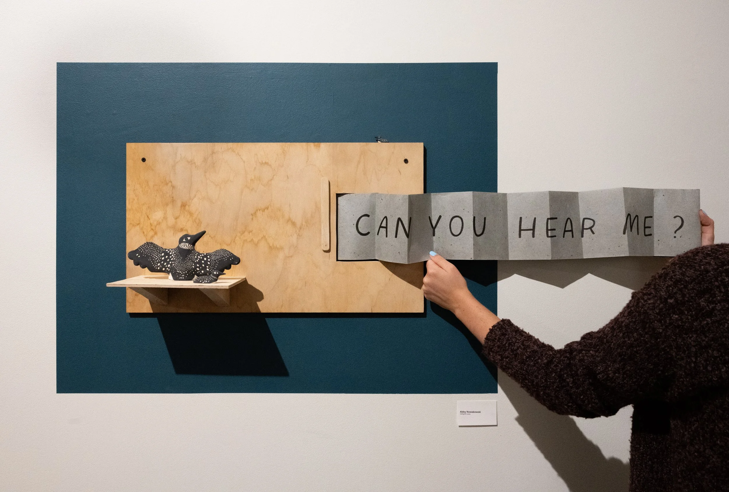 An art installation features a dark blue and wood-paneled background with a small, black, polka-dotted bird sculpture on a ledge. A person holds a folded gray paper sign with the handwritten message, "CAN YOU HEAR ME?"