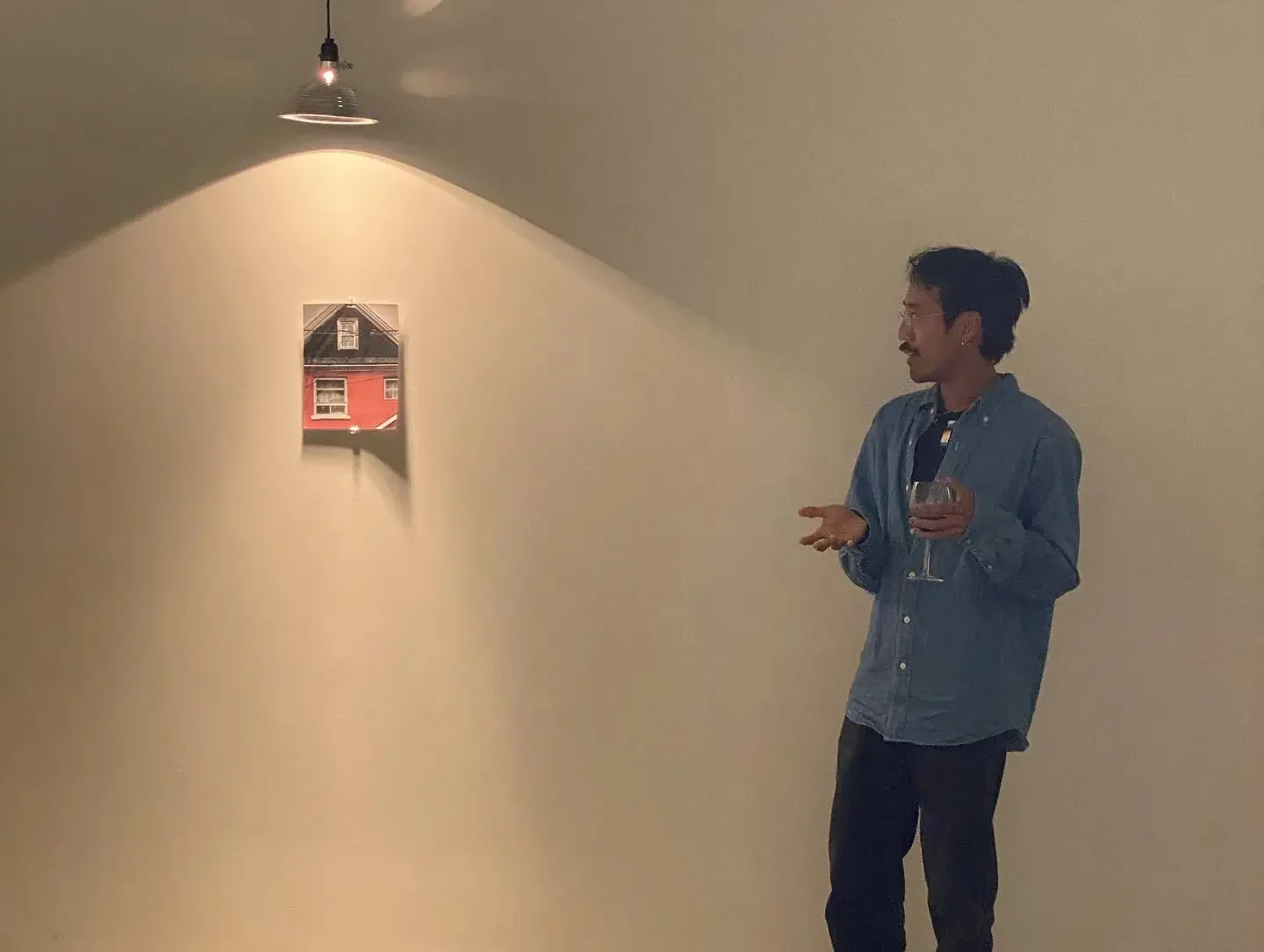 A man standing in a gallery or art space, holding a glass of wine, facing a small, illuminated artwork on the wall. The artwork depicts a building with a red facade and windows.