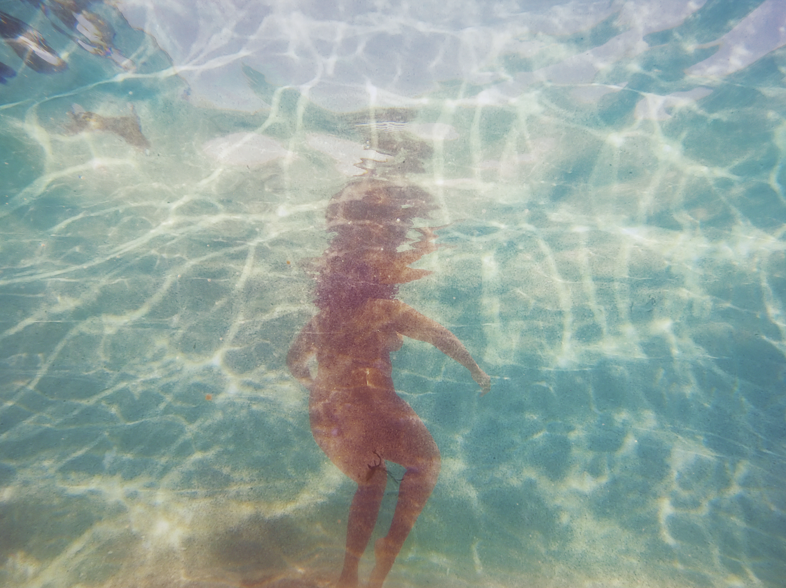 A person with long hair swimming underwater in a pool with clear blue water.