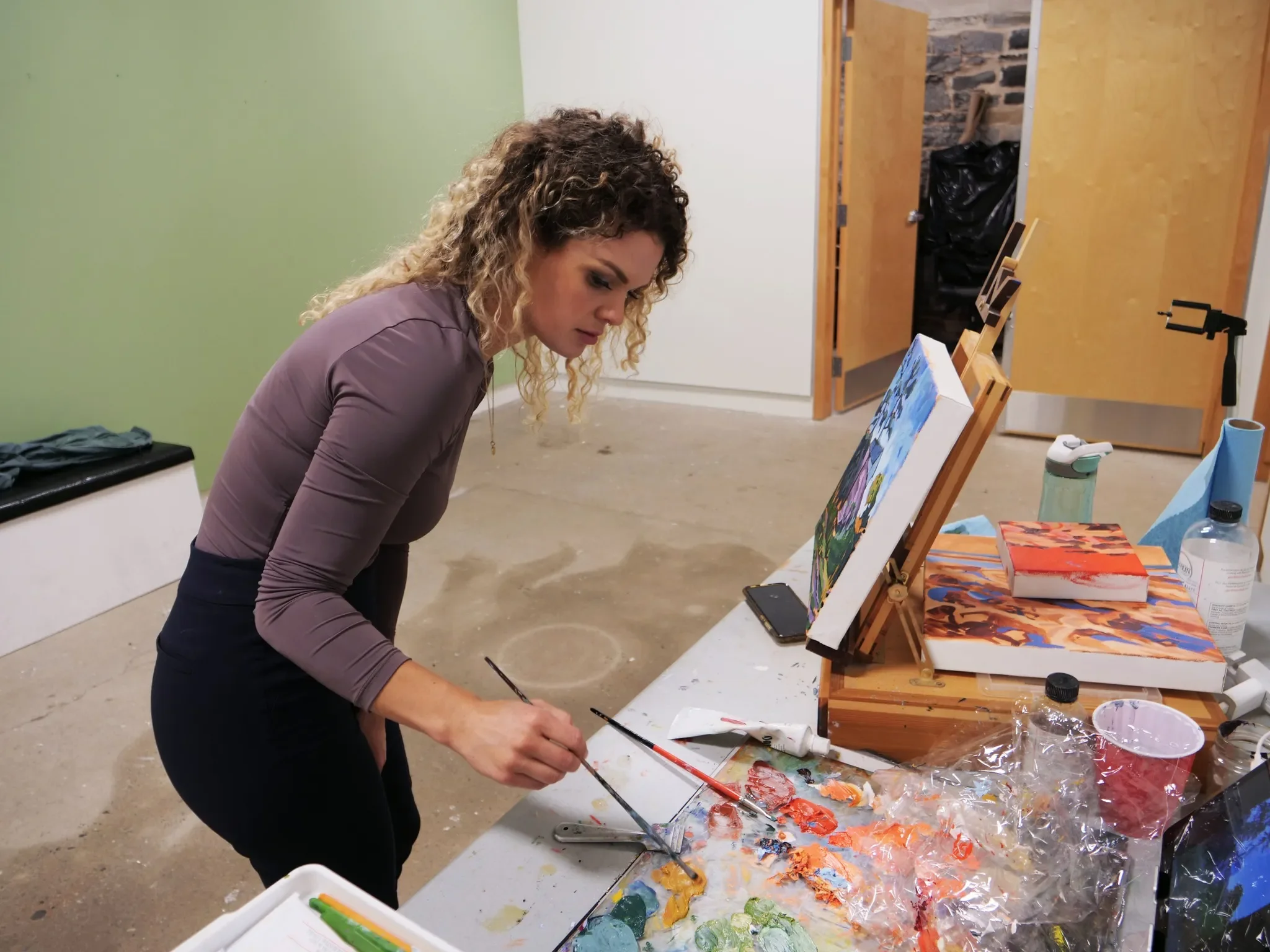 A woman with curly hair wearing a purple long-sleeve shirt and black pants, standing at a table in a studio, painting with braces of paint on a palette. The table holds art supplies, paintings on easels, and various paints and brushes, with a green w