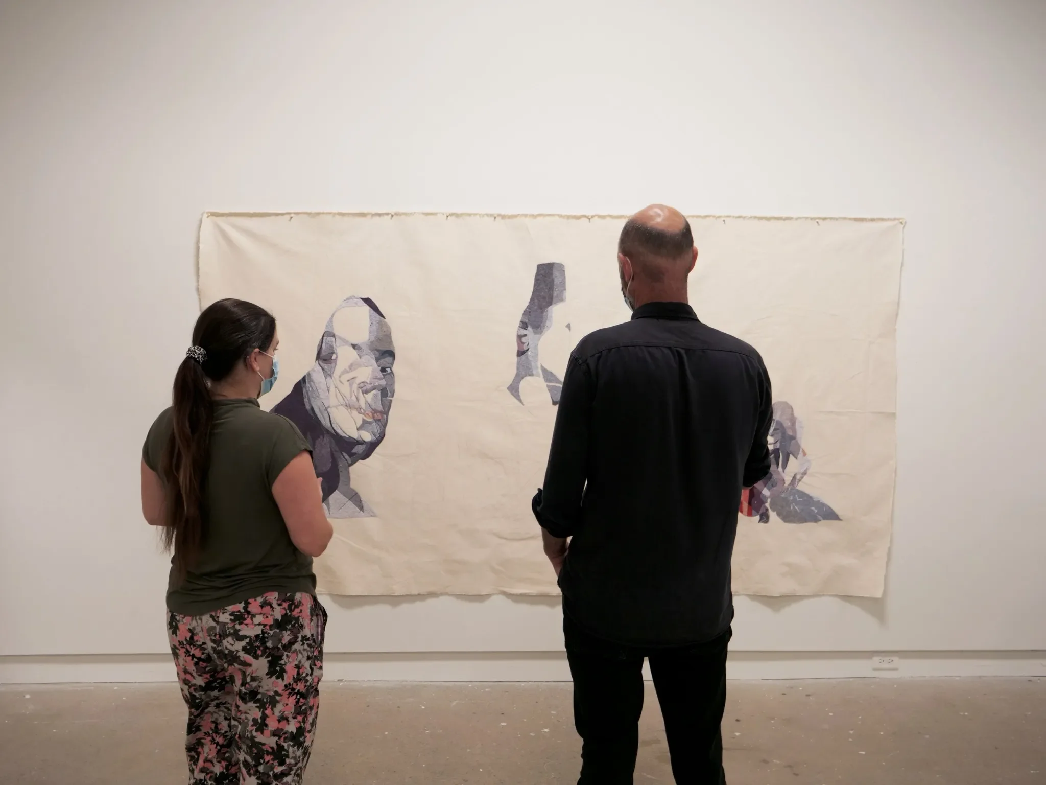 Two people wearing face masks observe a large textile artwork on a gallery wall, which features abstract portraits in a monochromatic palette.