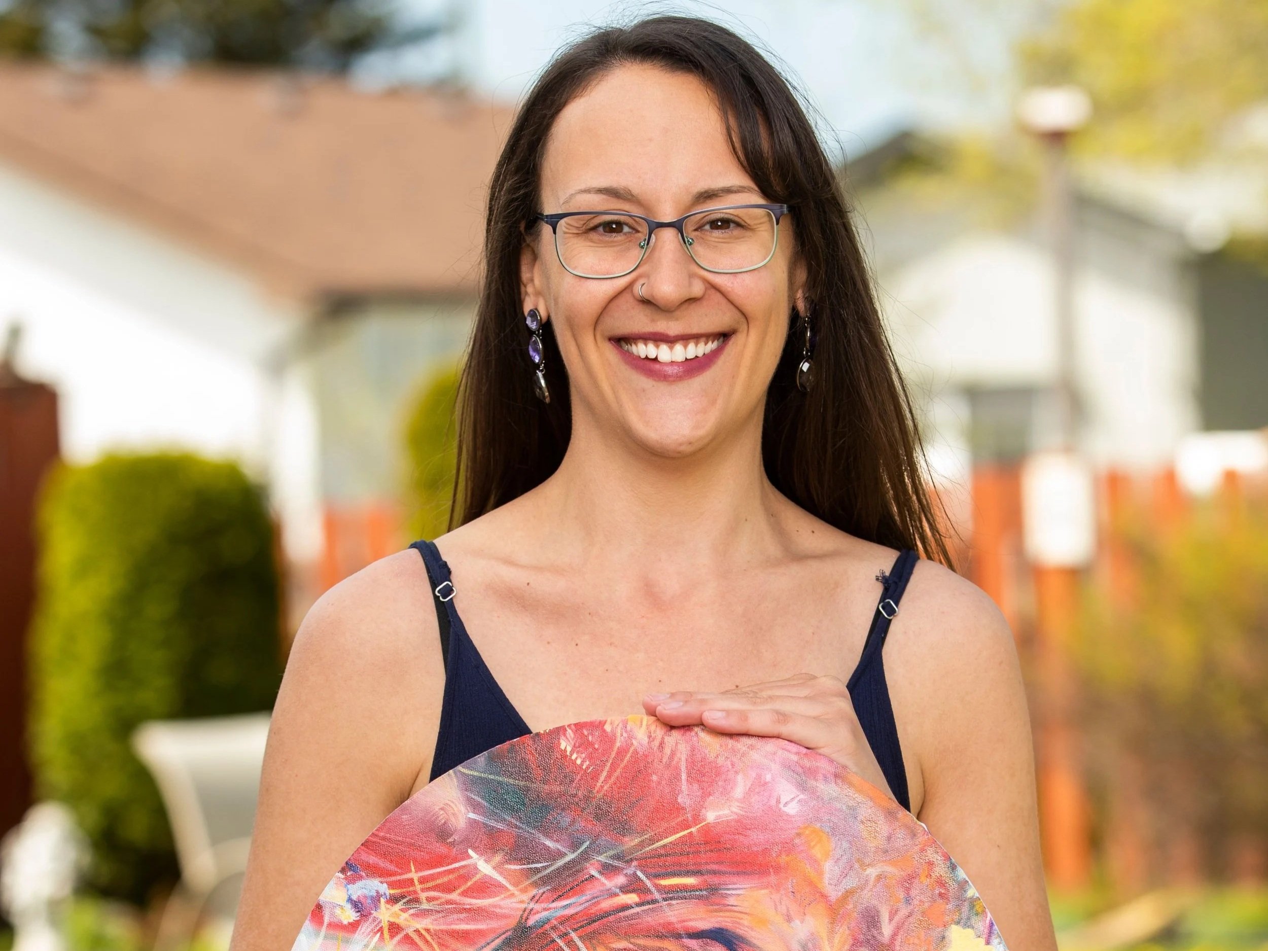 A woman with long dark hair, glasses, and earrings smiling outdoors, holding a colorful round fan.