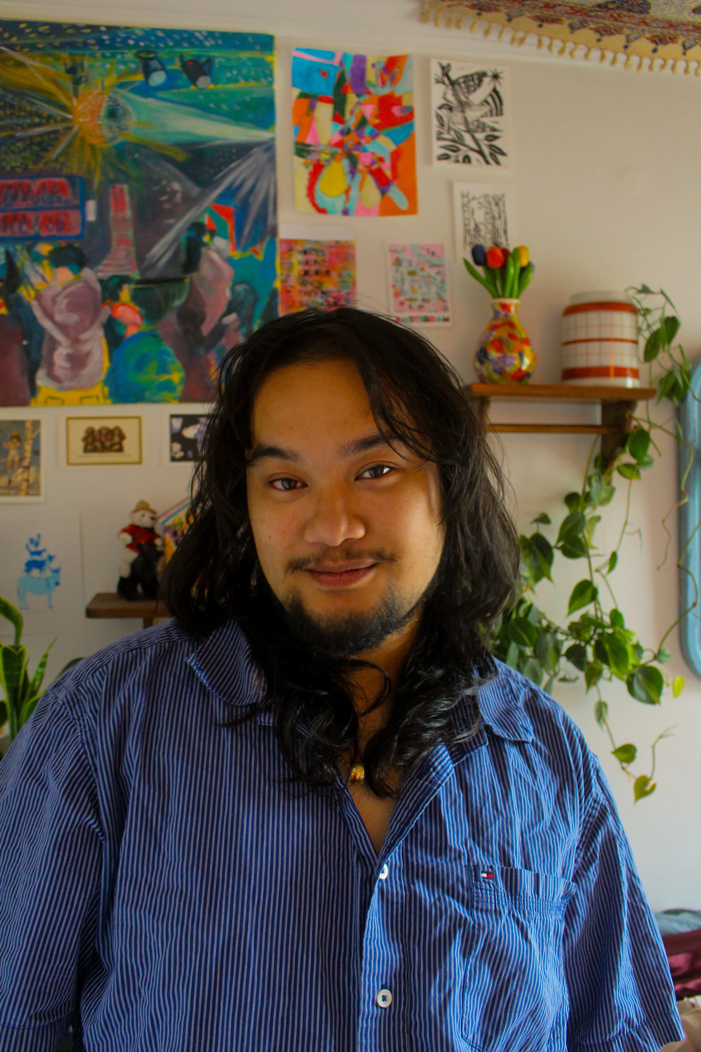 Portrait of a man with long black hair and a goatee, wearing a blue striped shirt, standing in a room with colorful artwork and plants in the background.