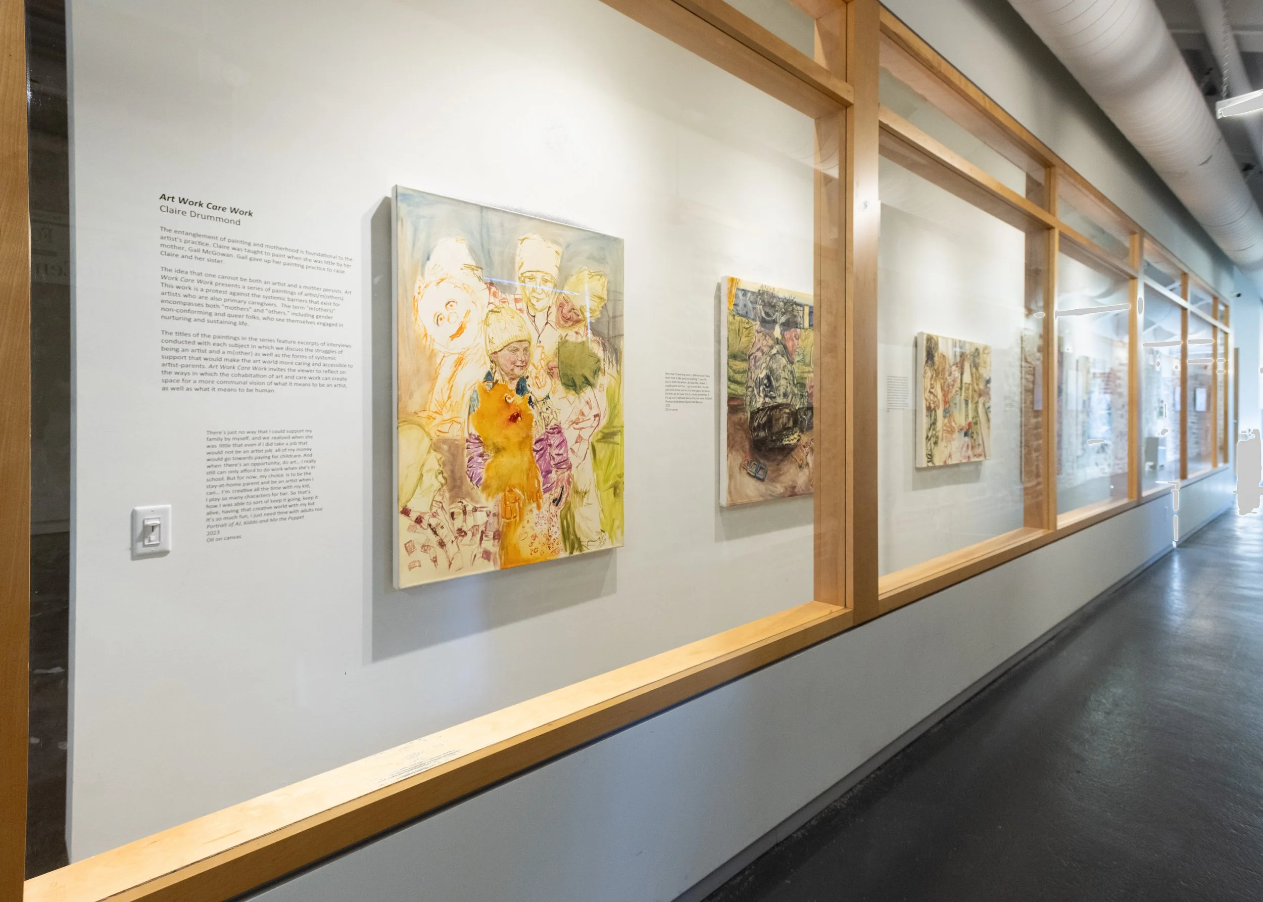 Art exhibition with paintings displayed on a white wall inside a gallery, wooden framing around the display area, and informational text about the artworks.