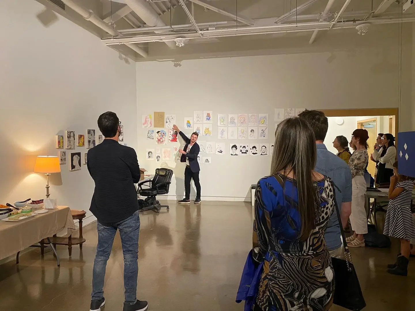 A group of people attending an art gallery viewing, with an exhibitor presenting and various artwork displayed on the walls.