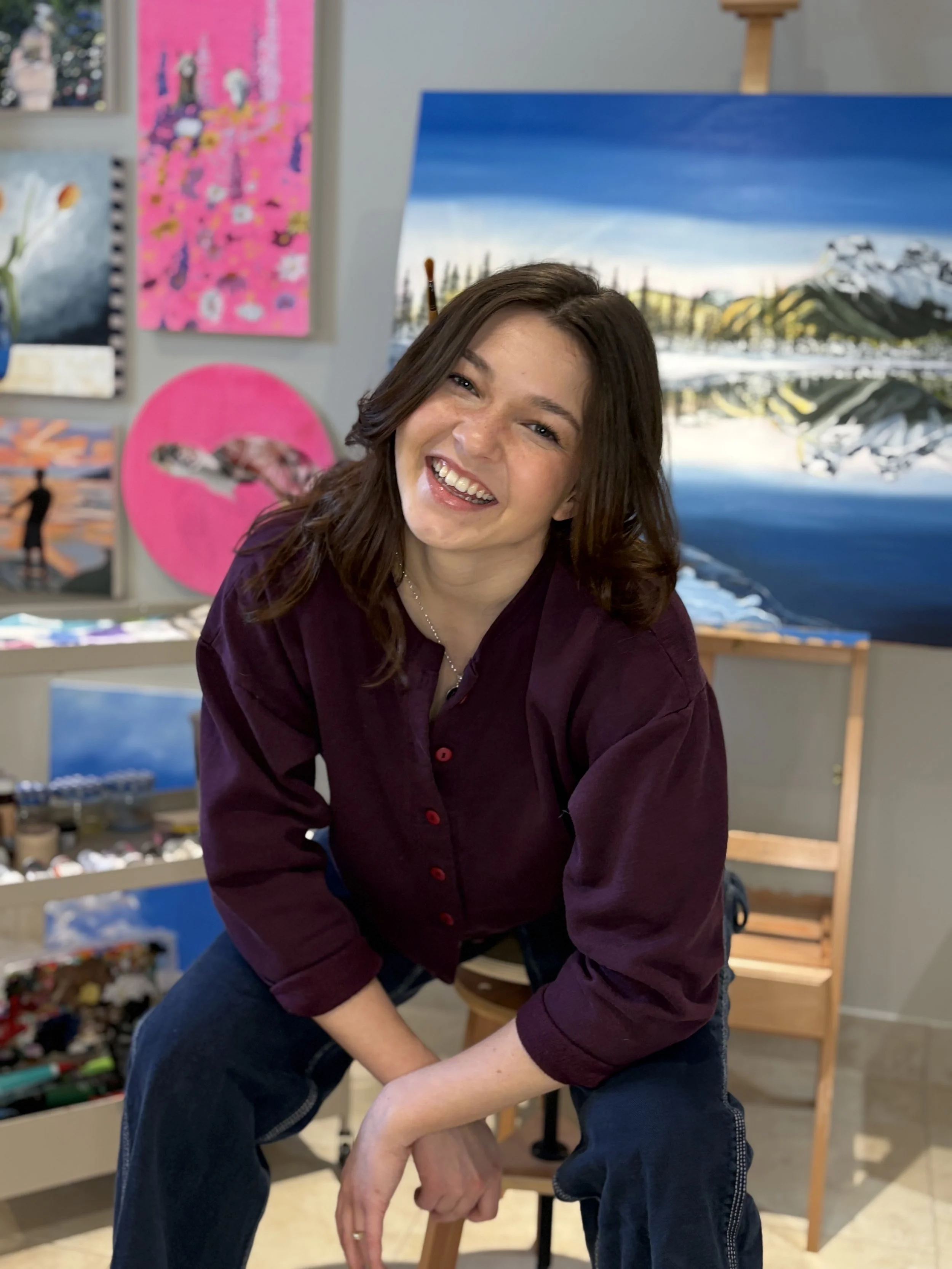 A young woman with shoulder-length brown hair, wearing a dark purple shirt and blue jeans, smiling and leaning forward in an art studio with paintings and art supplies in the background.