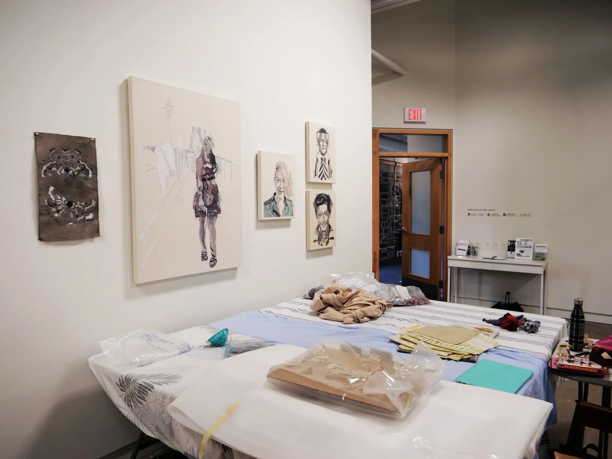 An art gallery or studio with a white wall featuring several paintings and sketches, a table covered with various art supplies, and a door leading to another room.