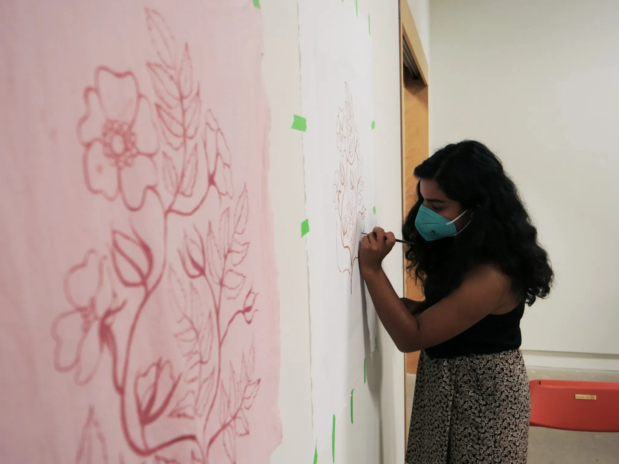 A woman with dark curly hair, wearing a teal face mask, a black sleeveless top, and patterned pants, is drawing on a large white paper attached to a wall with green tape.