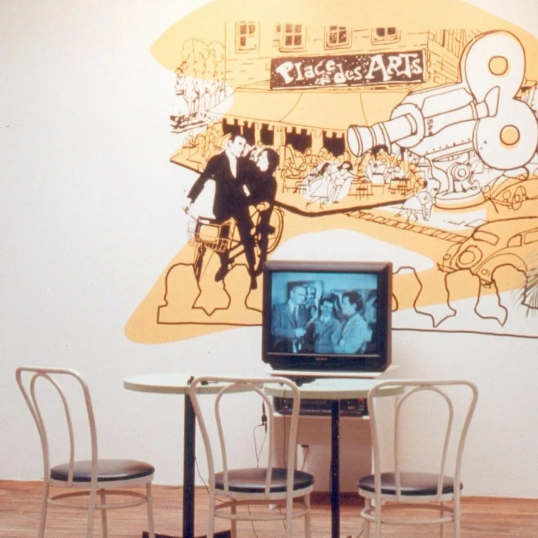 A small round table with three chairs in front of a wall decorated with a yellow and black mural. The mural depicts a city scene including a couple riding a bicycle, a large telescope, an audience at a plaza called 'Place des Arts,' and people sitting outdoors. A vintage television on a stand is on the table showing a black and white program.