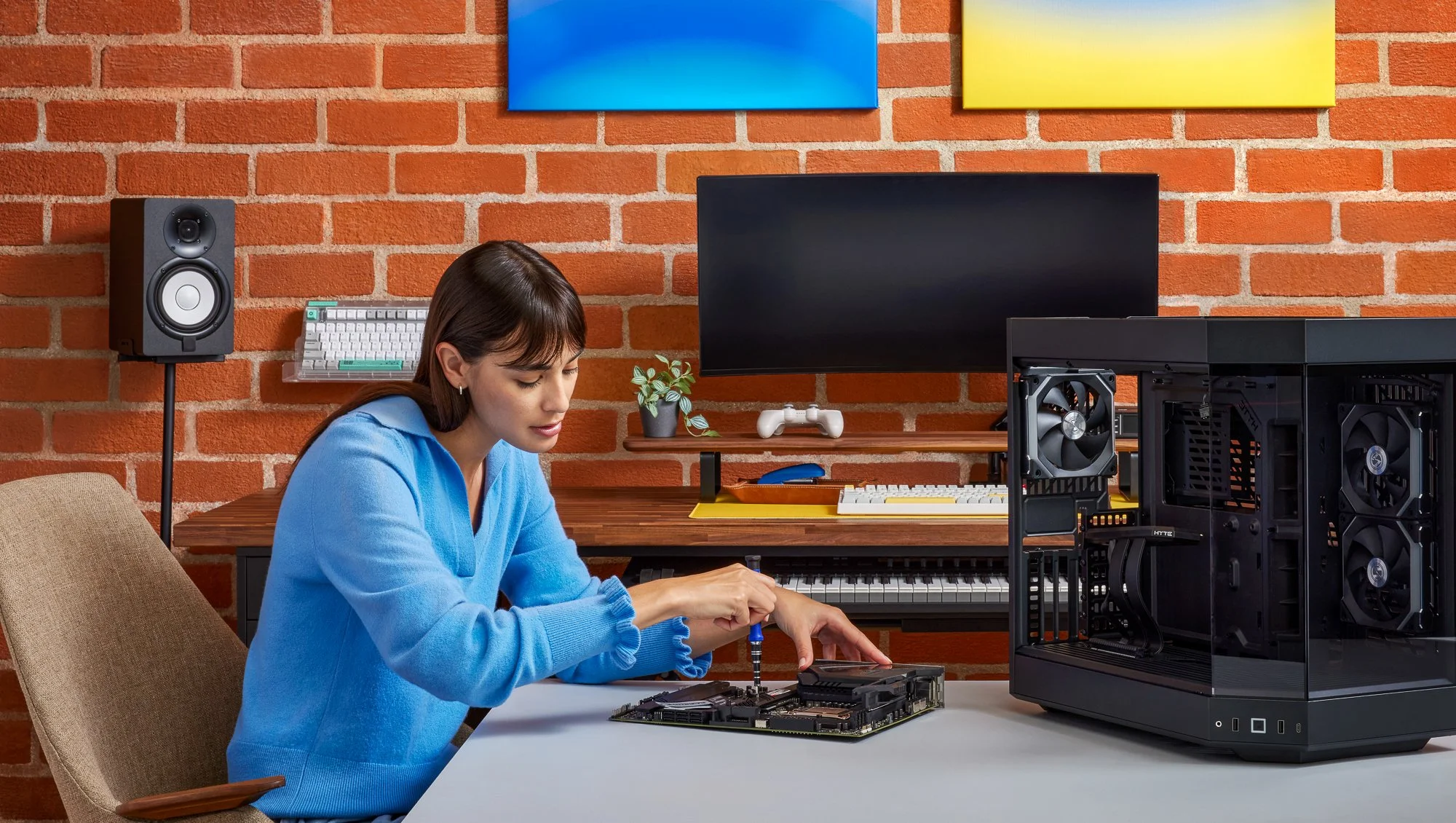 A woman assembling a computer inside a room with a brick wall. There is a computer tower case, a motherboard, and tools on the table. A monitor, speaker, keyboard, and a gaming controller are in the background.